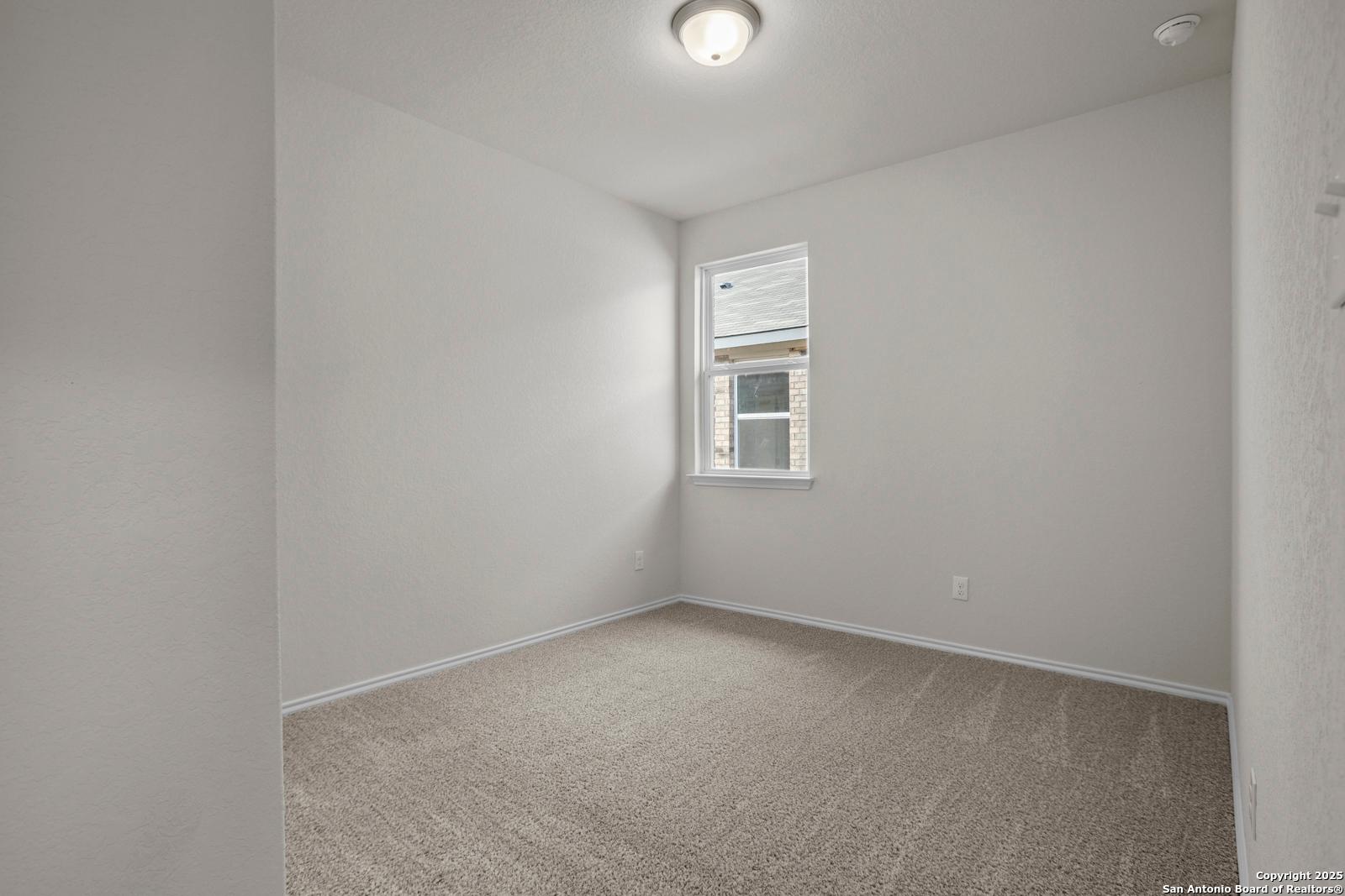Empty bedroom with neutral beige walls, tan carpet flooring, window AC unit, and flush ceiling light in Davidson Homes The Asheville J, San Antonio, TX