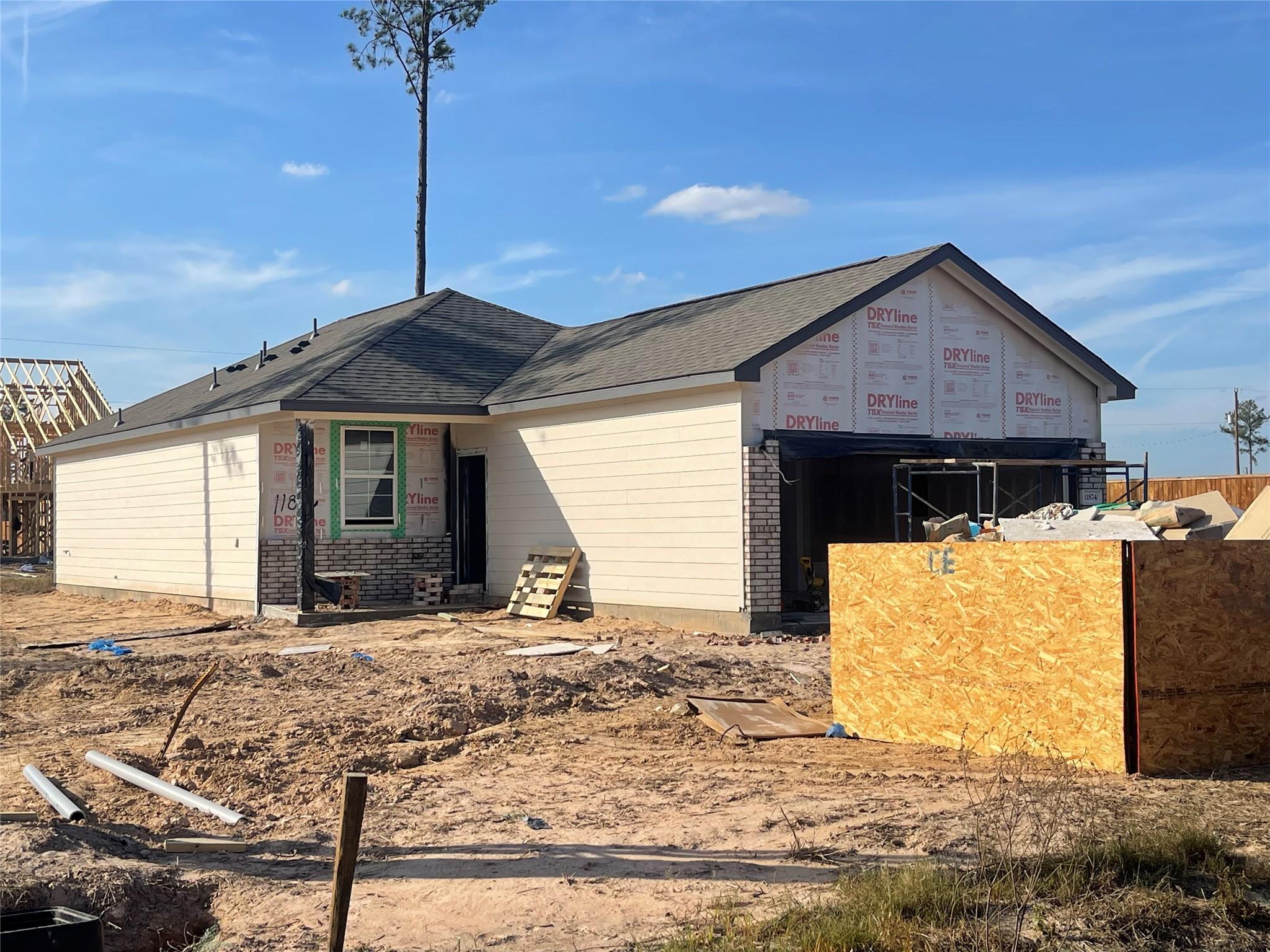 Under-construction single-story 3-bedroom home with beige siding, dark shingled roof, and open 2-car garage in Spring Branch Crossing, Conroe, Texas