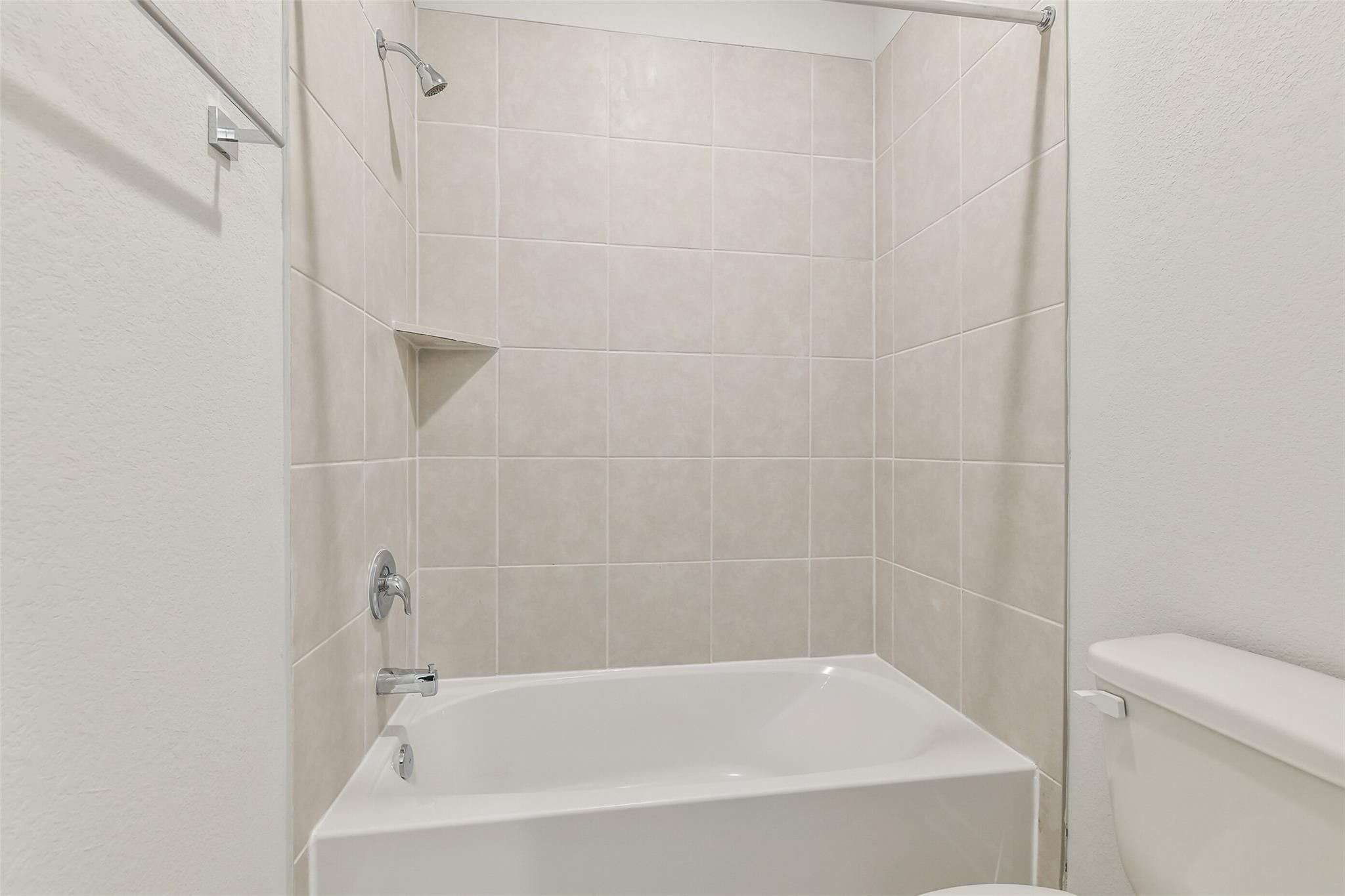Modern bathroom with white bathtub-shower combo, beige tile walls, niche shelf, and toilet in Davidson Homes The Colorado F, Cleveland, Texas