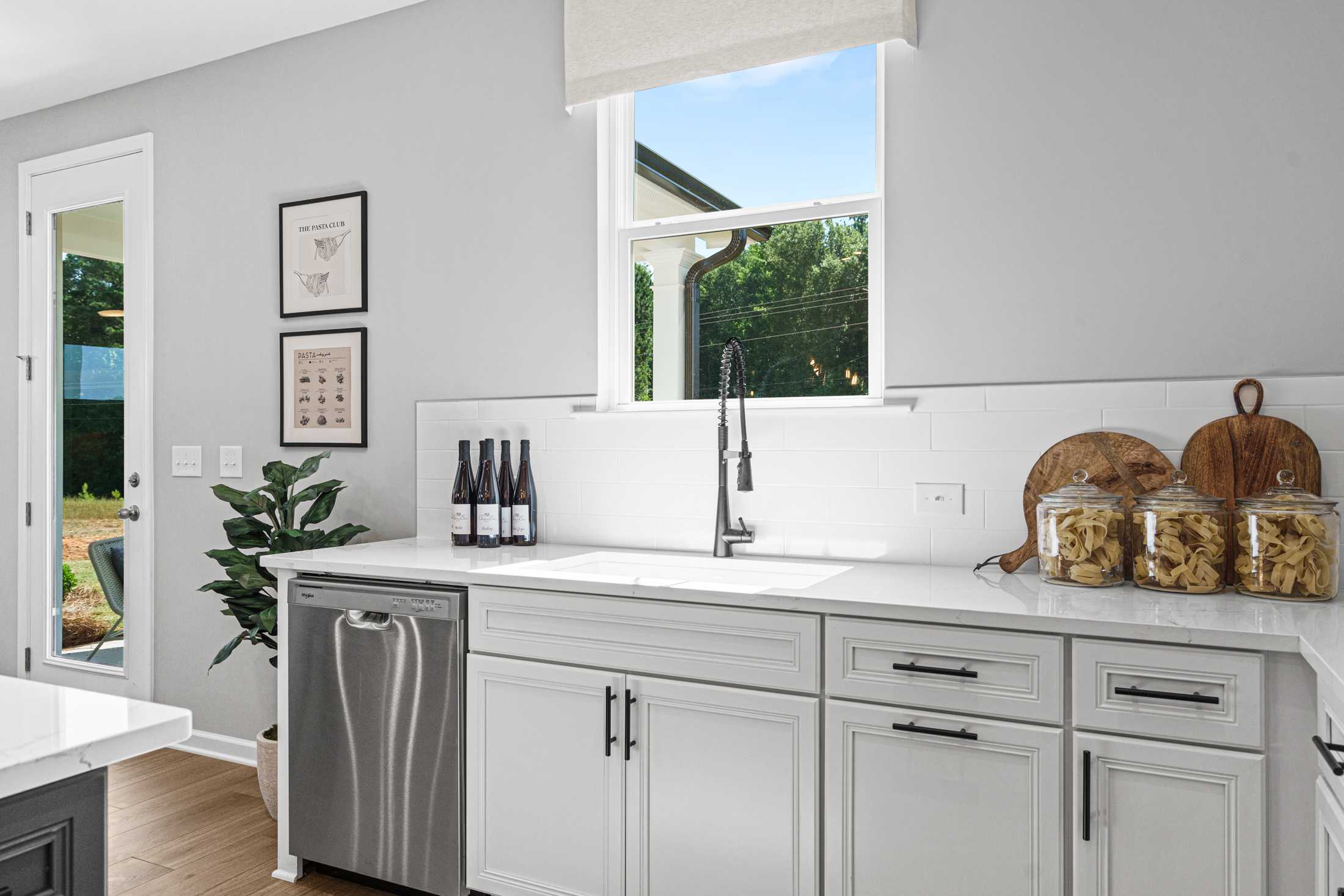 Modern kitchen in The Hickory B showcasing white shaker cabinets, stainless steel sink, wine bottles, and sunny garden window