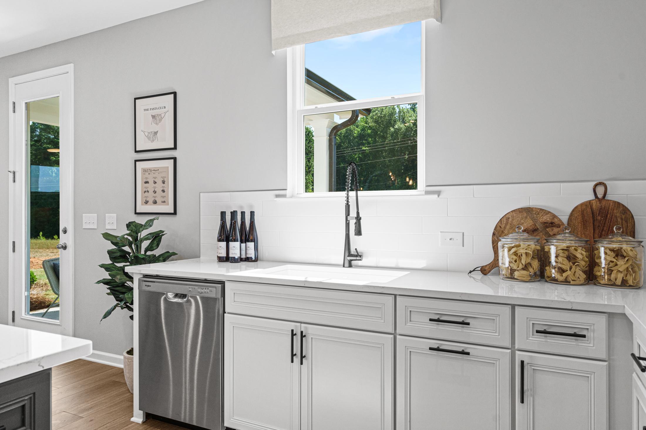 Modern kitchen in The Hickory B showcasing white shaker cabinets, stainless steel sink, wine bottles, and sunny garden window