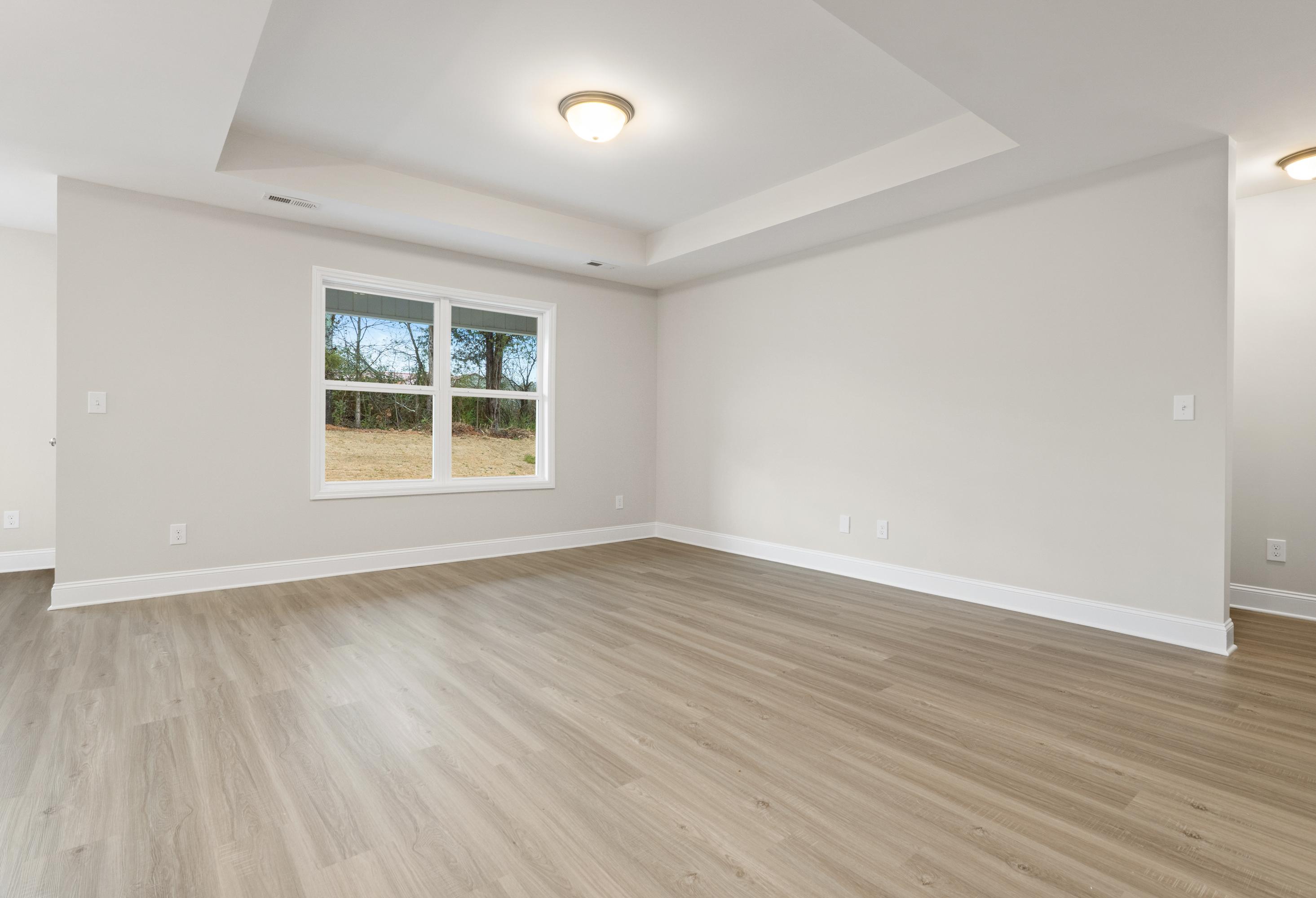 Spacious empty master bedroom in The Asheville with tray ceiling, large window, beige walls, and luxury vinyl plank flooring