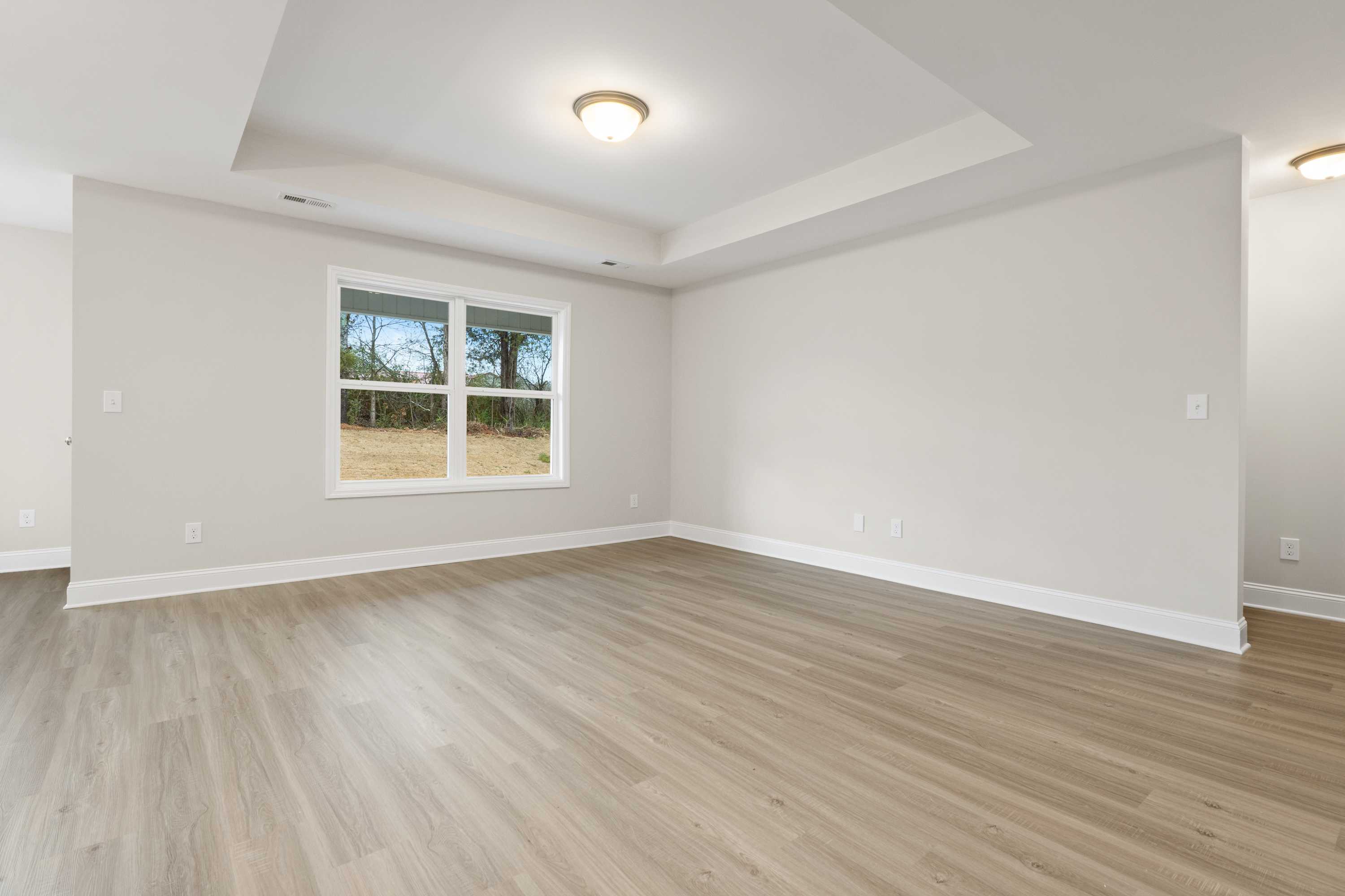 Spacious master bedroom in The Asheville home design with light oak floors, beige walls, and large window overlooking trees