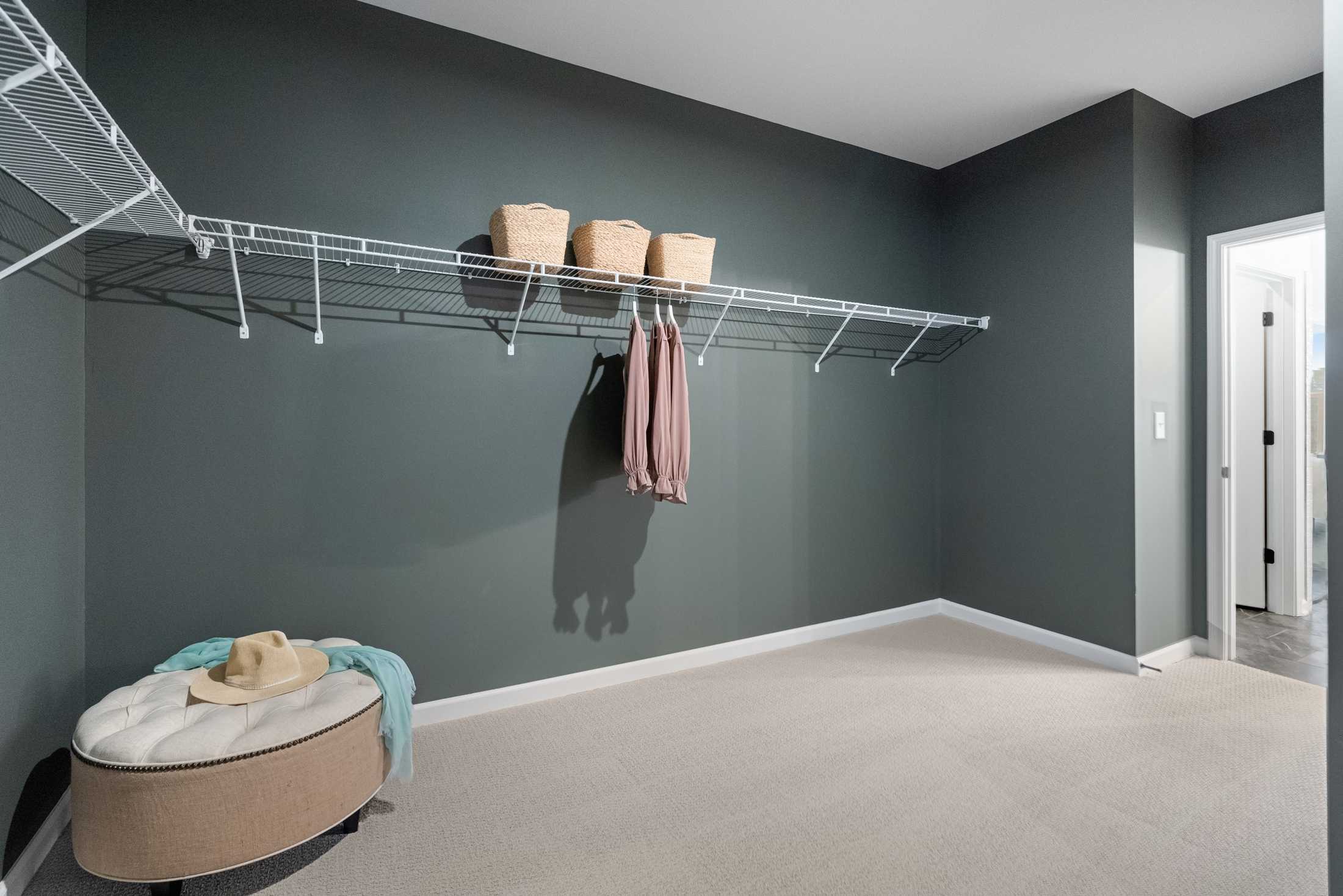 Spacious walk-in closet in The Edison C master suite with white wire shelving, gray walls, hanging blouse, storage baskets, and upholstered ottoman