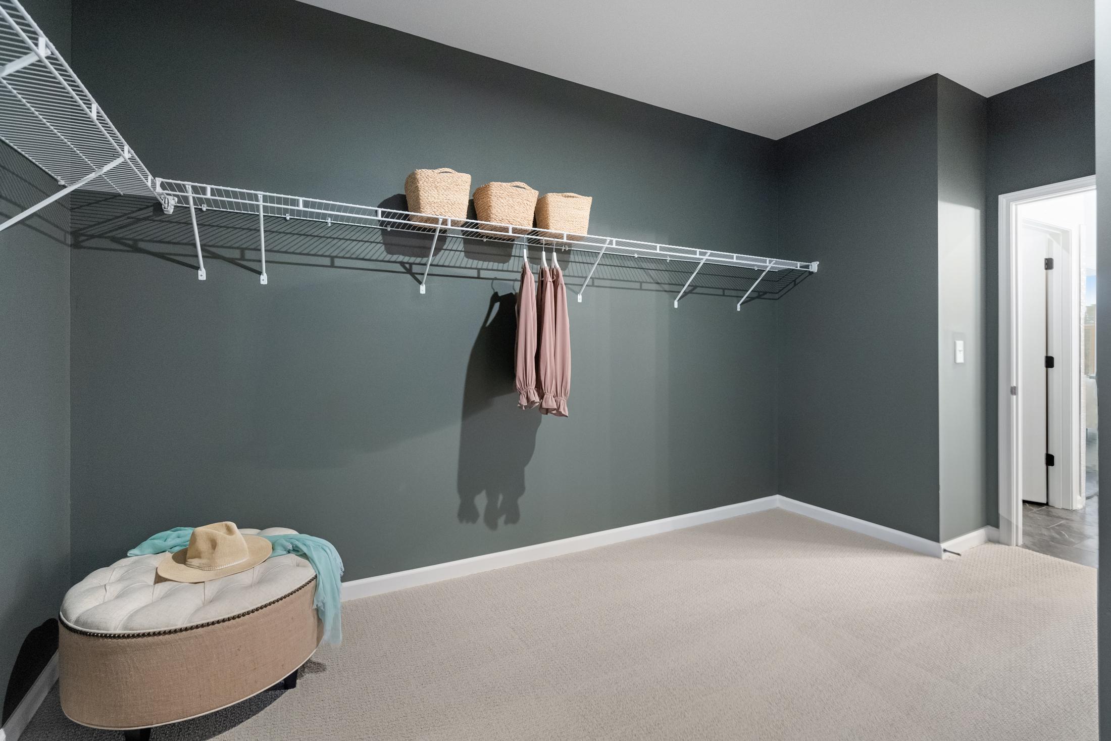 Spacious walk-in closet in The Edison A master suite featuring white wire shelving, gray walls, storage baskets, hanging blouse, and upholstered ottoman