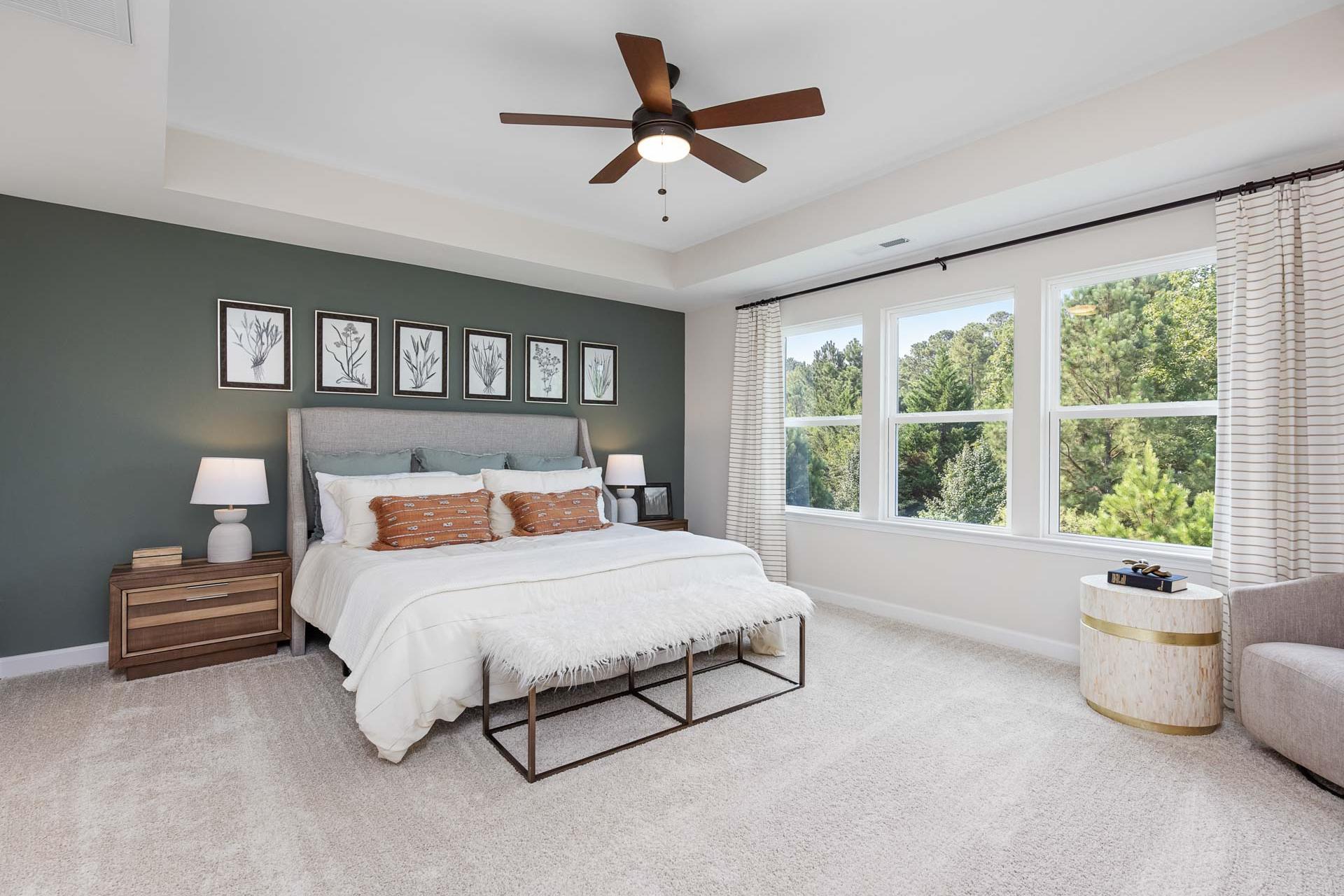 Spacious primary bedroom in Sierra Heights Clayton NC with king bed, gray accent wall, botanical prints, and large tree-view windows