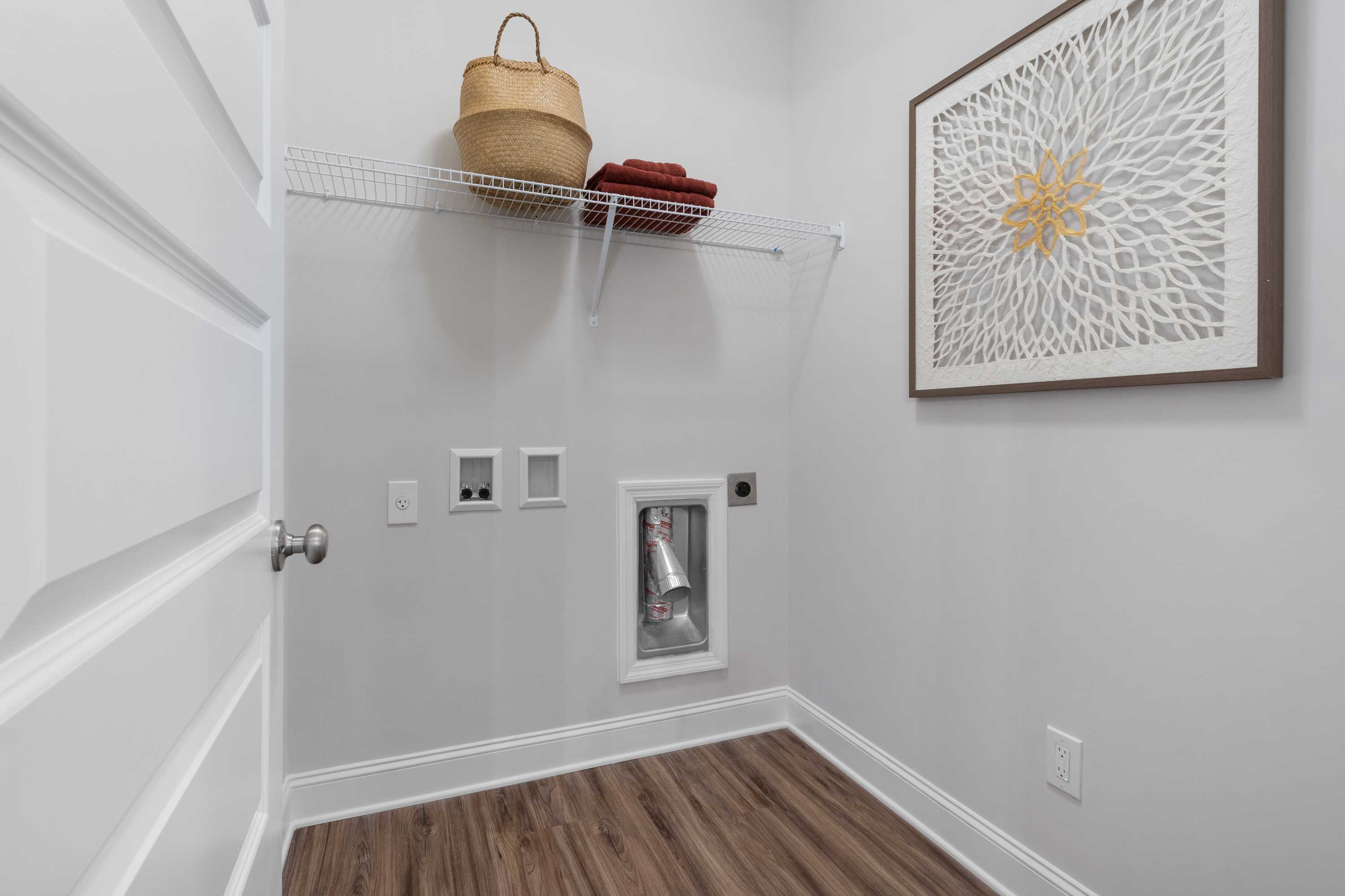 Laundry closet at The Retreat at Cain Park in Hartselle AL featuring shelves with wicker basket, washer dryer hookups, framed floral art, hardwood floors