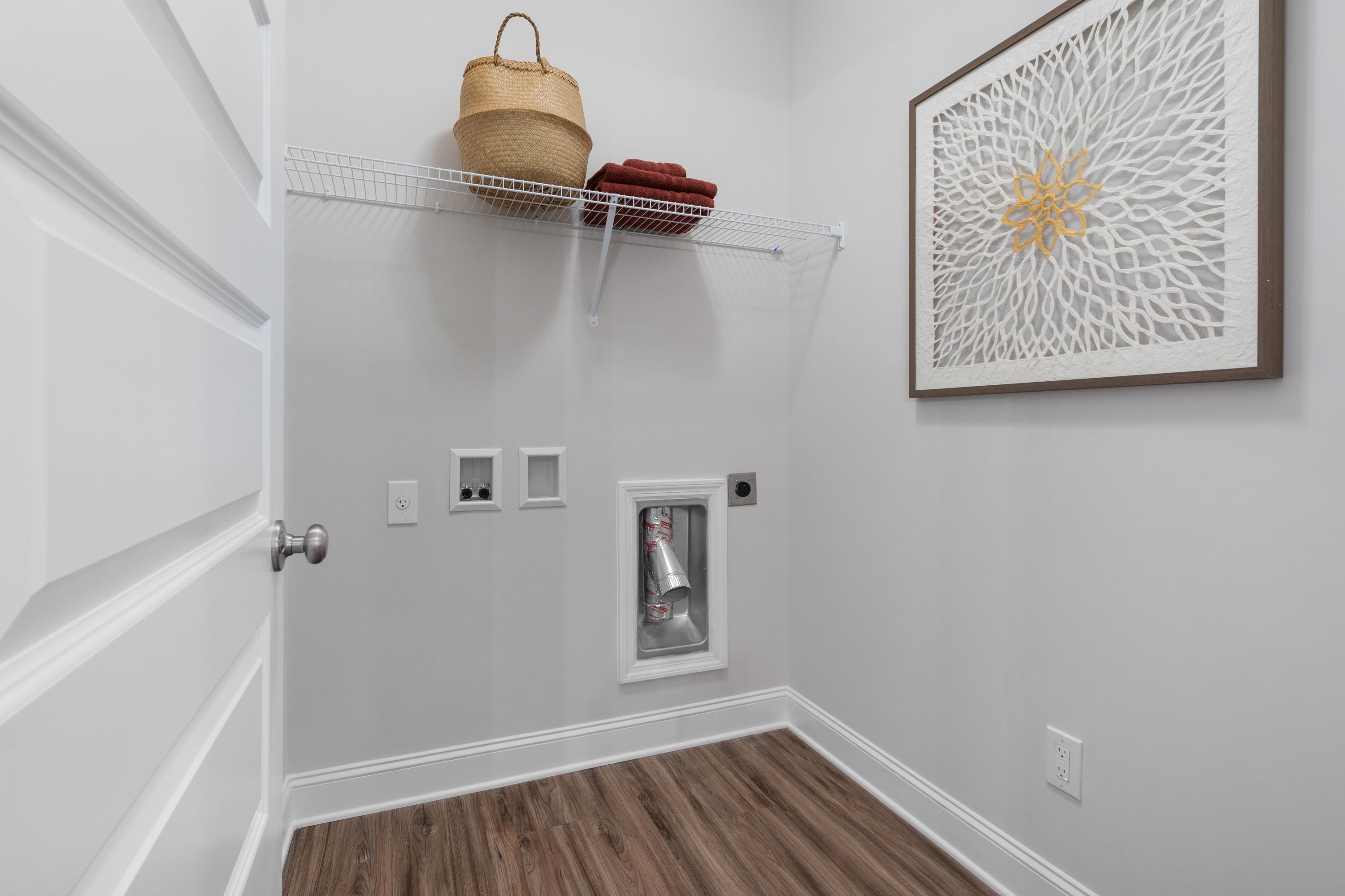 Laundry closet at The Retreat at Cain Park in Hartselle AL featuring shelves with wicker basket, washer dryer hookups, framed floral art, hardwood floors