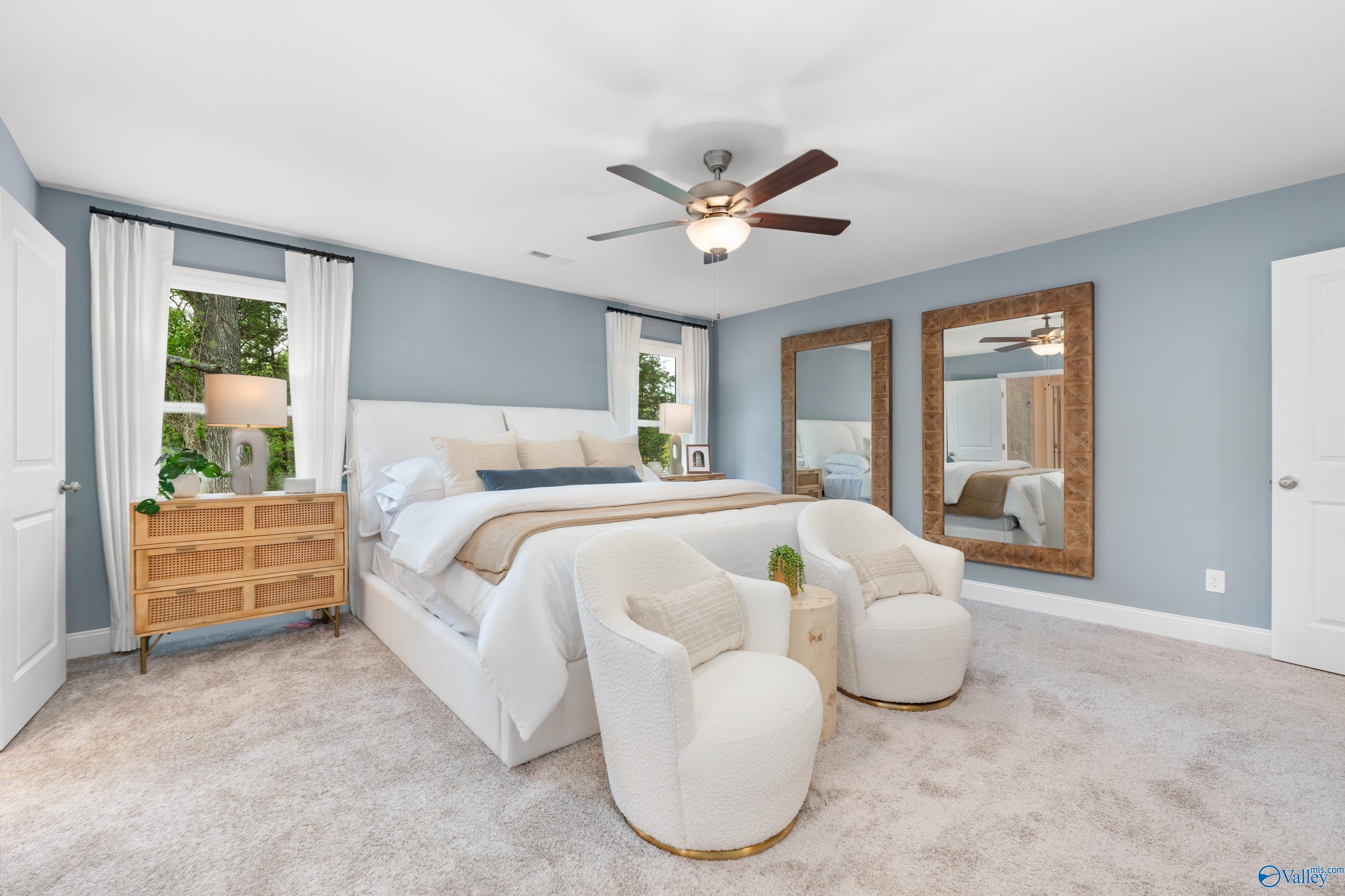 Serene master bedroom with white king bed, tufted armchairs, wooden dresser, large mirrors, and ceiling fan in Davidson Homes The Camden, Huntsville AL