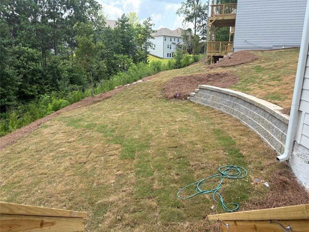 Sloped backyard with curved beige retaining wall, patchy grass, and pine straw mulch beside 5-bedroom Davidson Homes in Riverwood, Dallas, Georgia