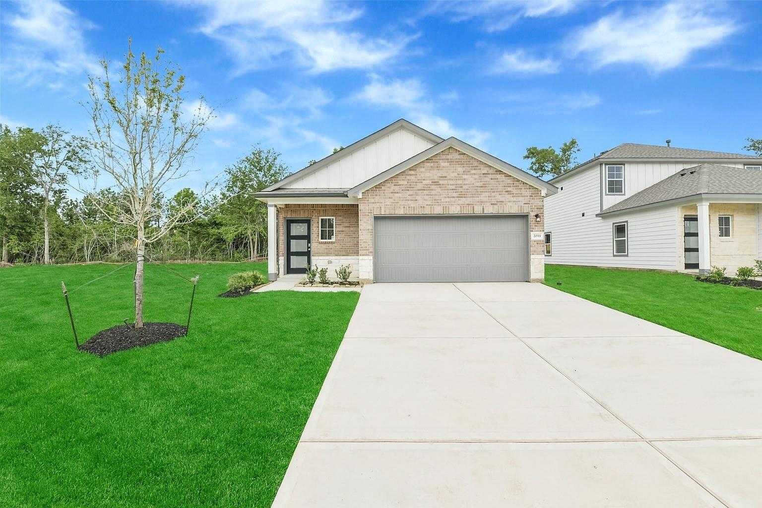 Modern tan brick 1-story home with 2-car garage, front porch, and lush green lawn in Caney Creek Place, Conroe, Texas