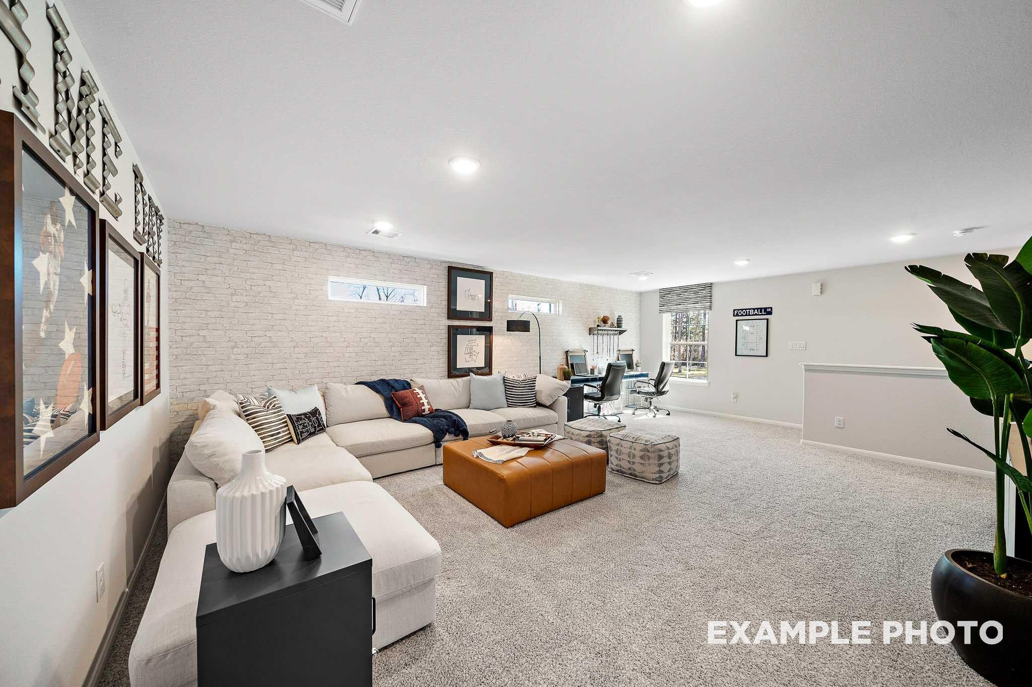 Spacious bonus room in The San Marcos home featuring L-shaped beige sofa, leather ottoman, desk workspace, and potted plant