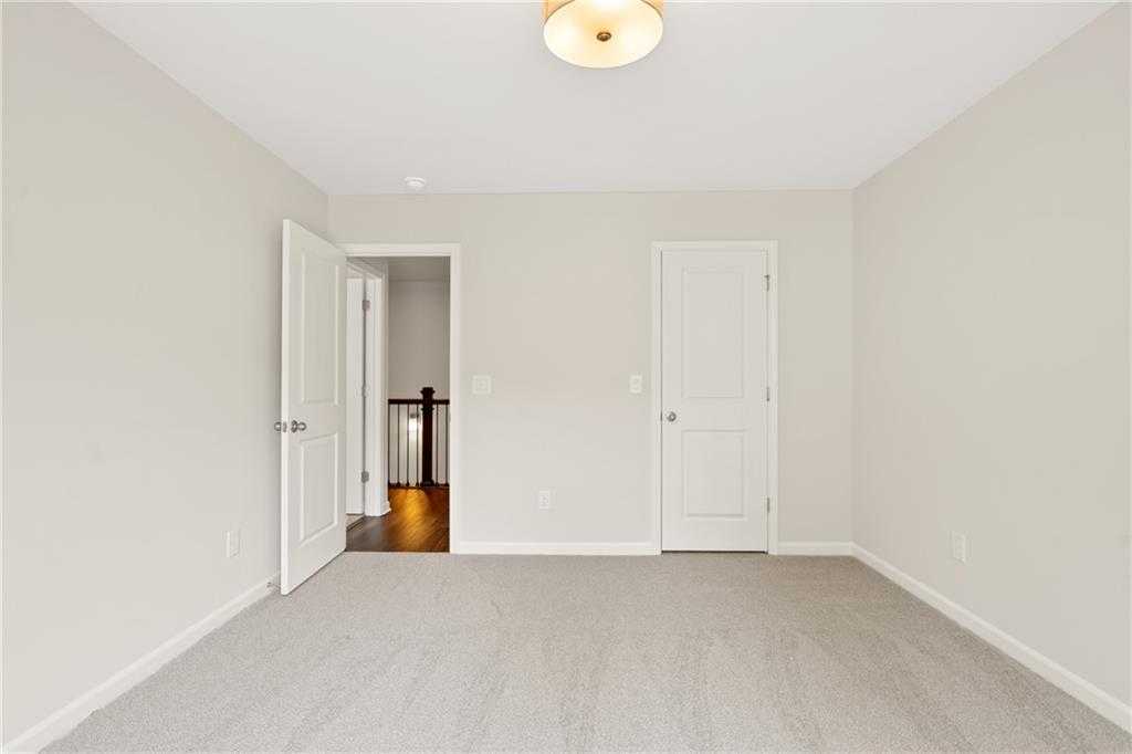 Bright empty bedroom with beige walls, gray carpet, and open door to staircase hallway in Davidson Homes The Cary B, Kennesaw, Georgia