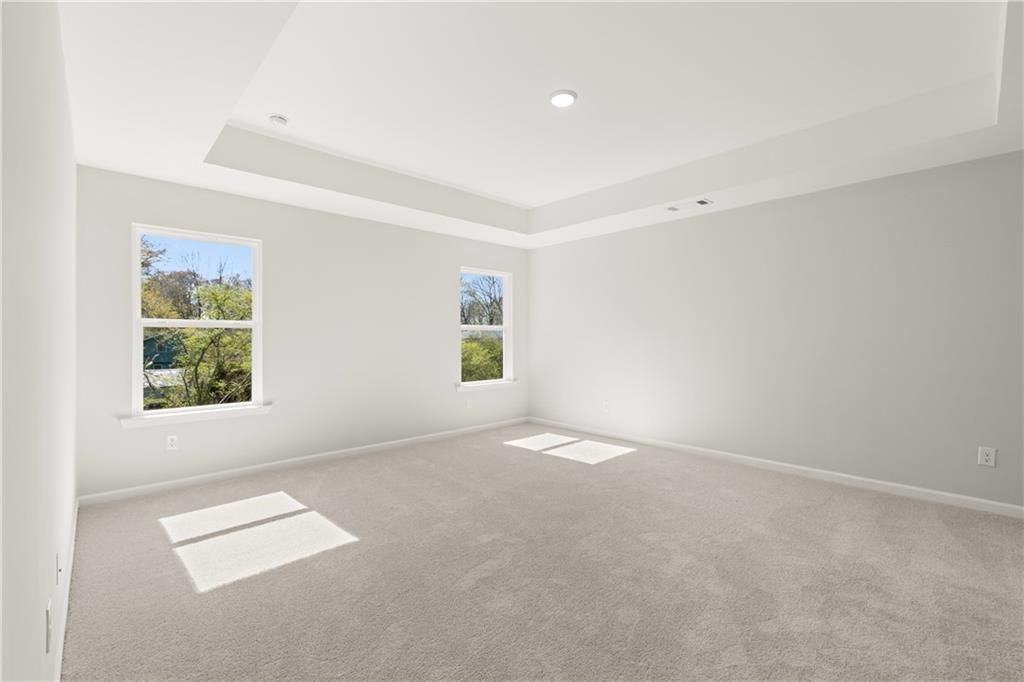 Bright empty bedroom with large windows, gray walls, tray ceiling, and beige carpet in The Hickory B, Winder, Georgia