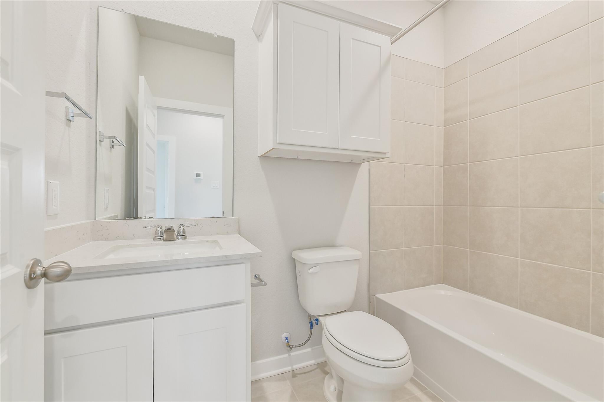 Modern guest bathroom featuring white vanity, quartz sink, toilet, bathtub, and beige tiled walls in The Frio G by Davidson Homes, Conroe, TX