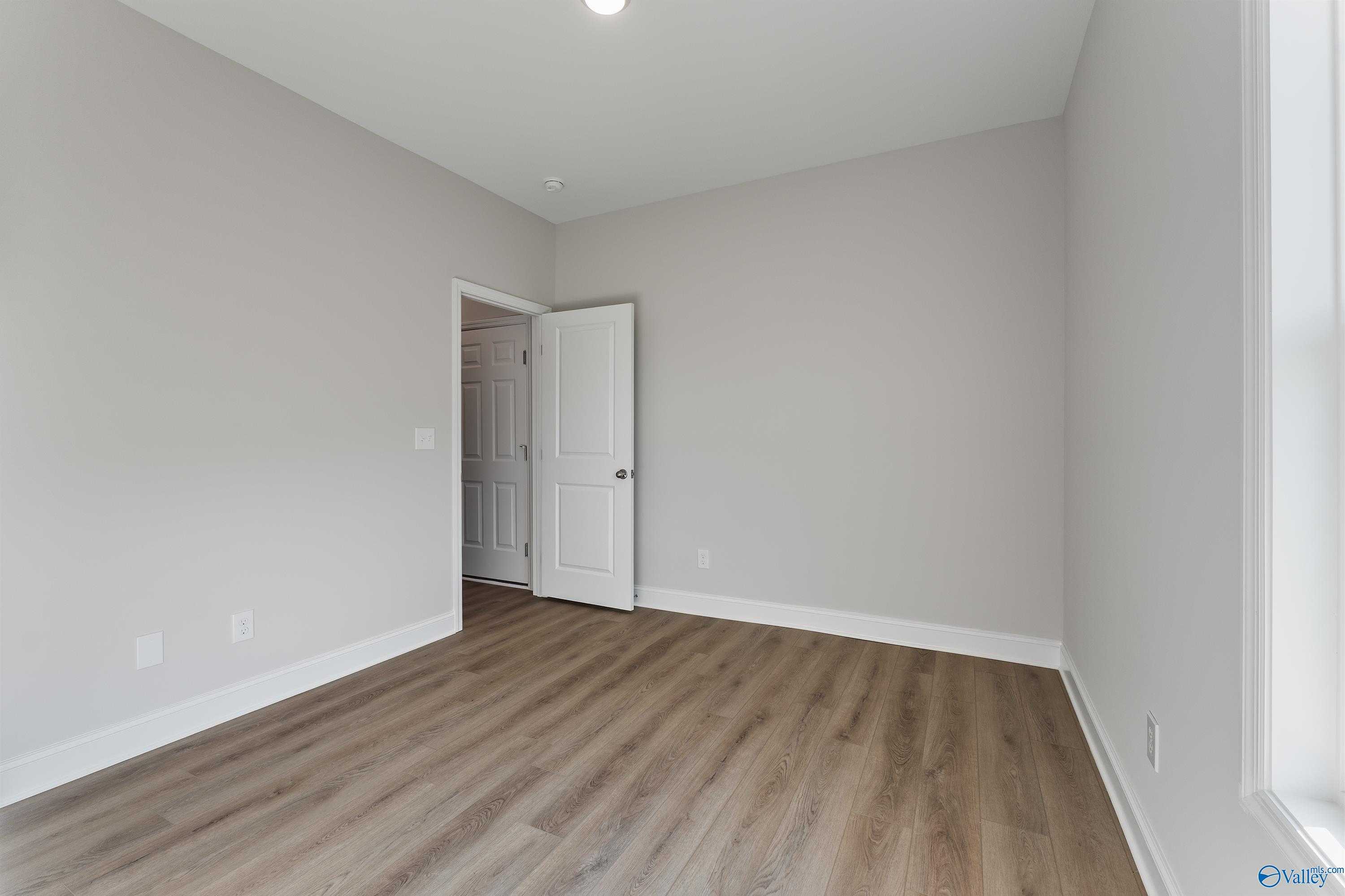 Spacious empty bedroom with light gray walls, white door, and laminate wood flooring in Davidson Homes The Daphne, Hazel Green, Alabama