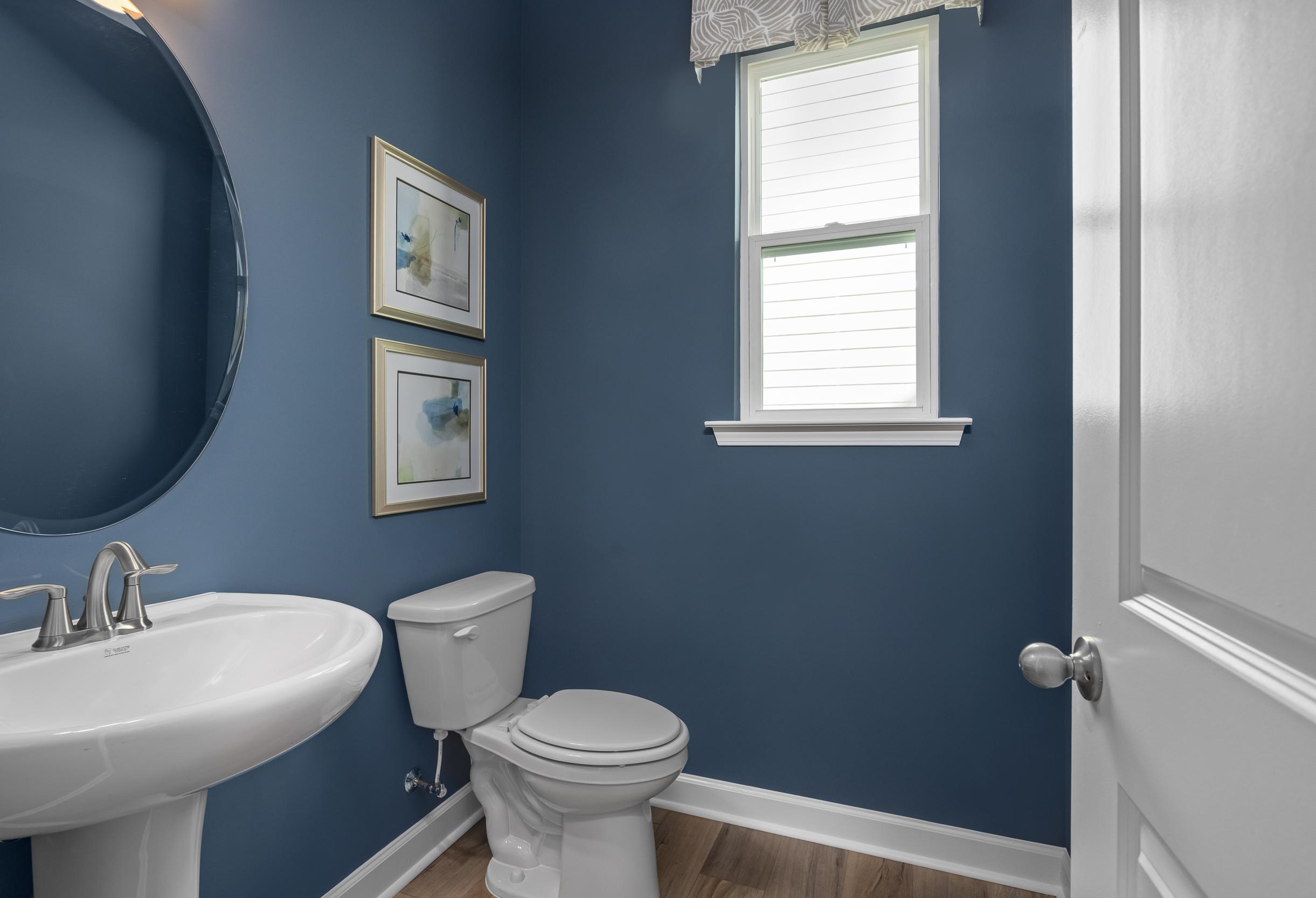 Cozy powder room in The Hickory II A with navy blue walls, pedestal sink, round mirror, and window accents