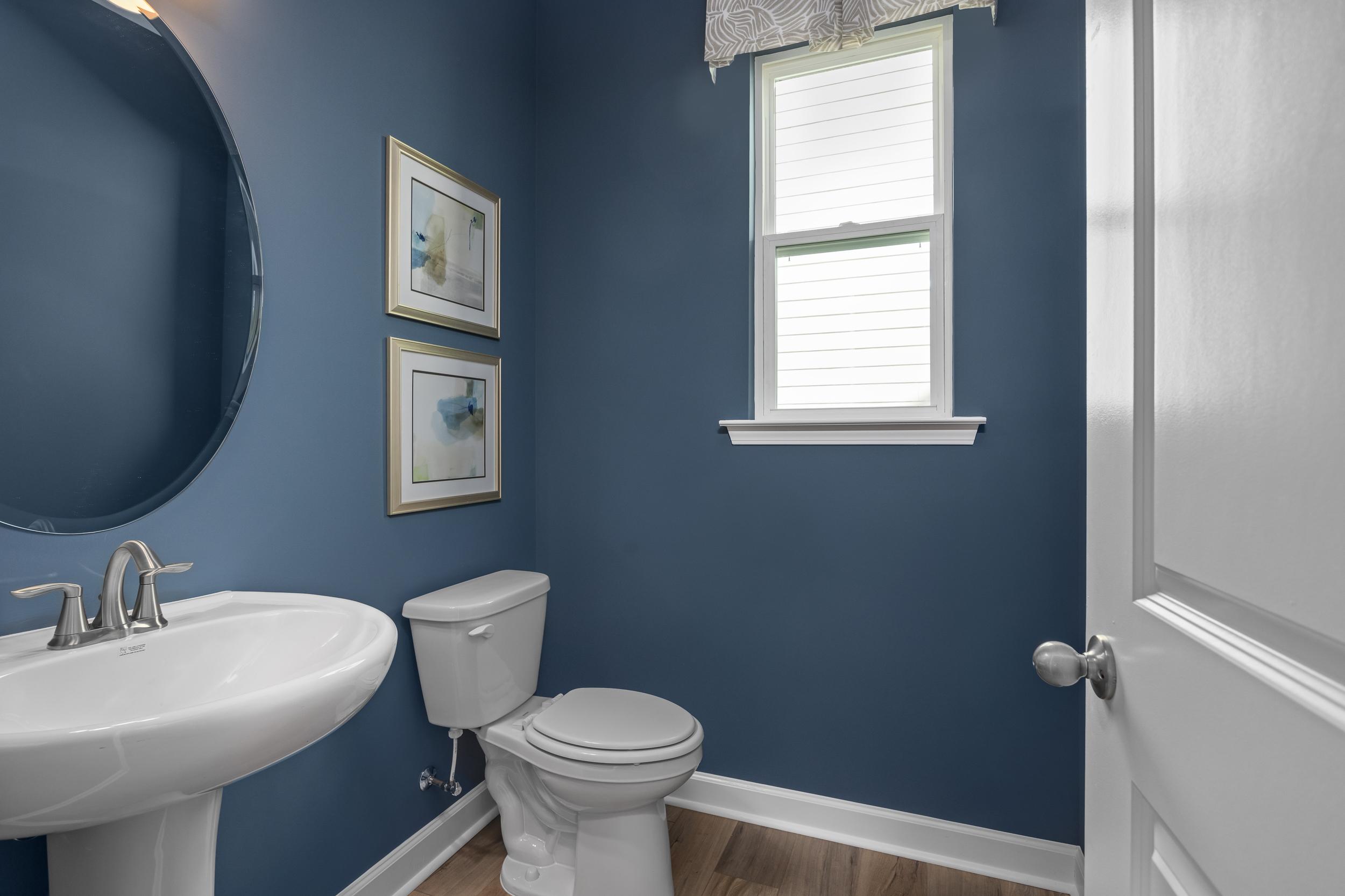 Cozy navy blue powder room in The Hickory II featuring pedestal sink, toilet, round mirror, and framed artwork