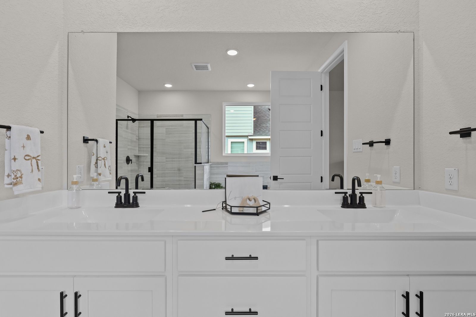 Modern master bathroom featuring double white vanity, black faucets, frameless mirror, and glass shower in Davidson Homes The Lanier G, Castroville, Texas
