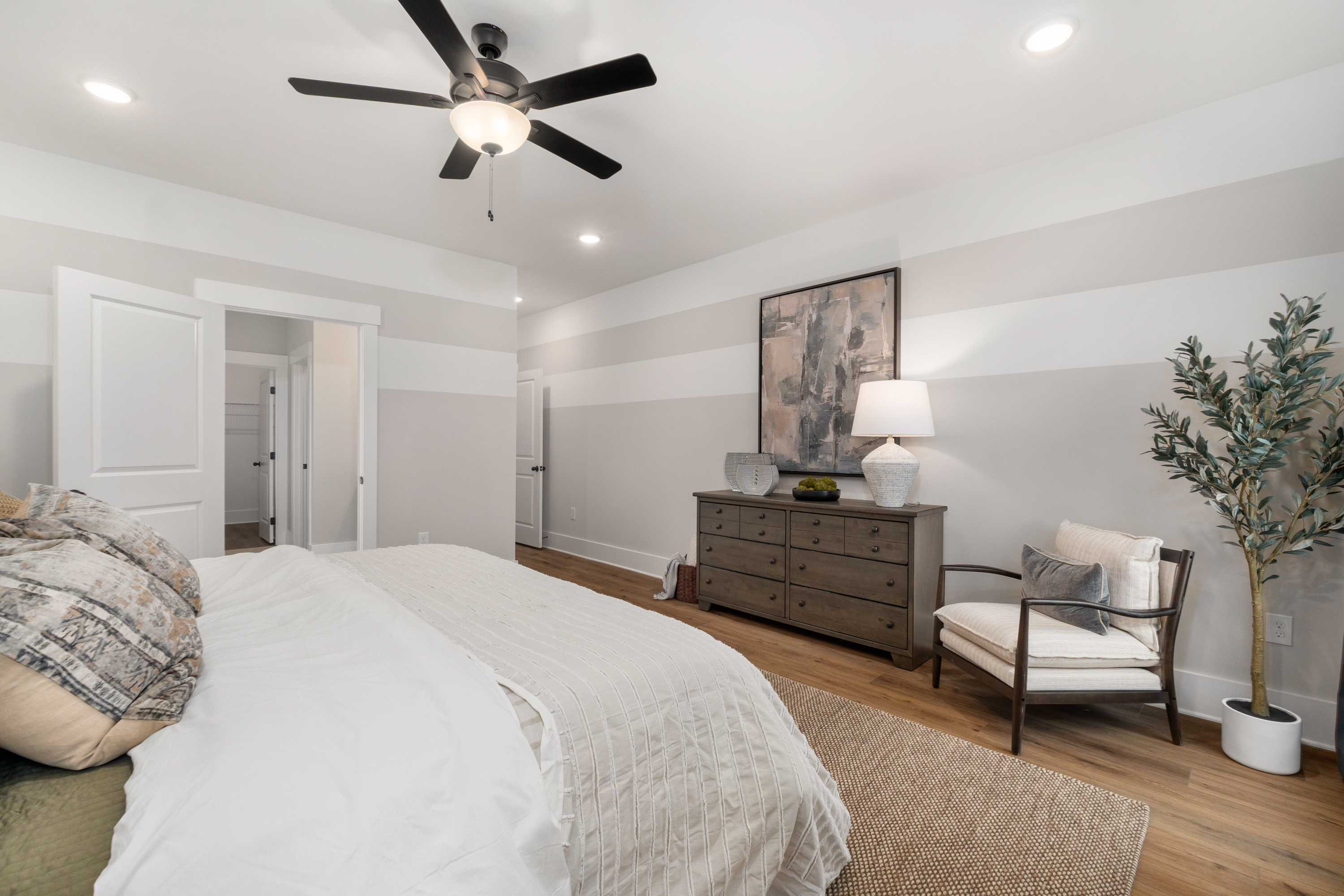 Cozy bedroom at Kendall Farms in Toney Alabama featuring ceiling fan, king bed with beige linens, dresser, armchair, and hardwood floors