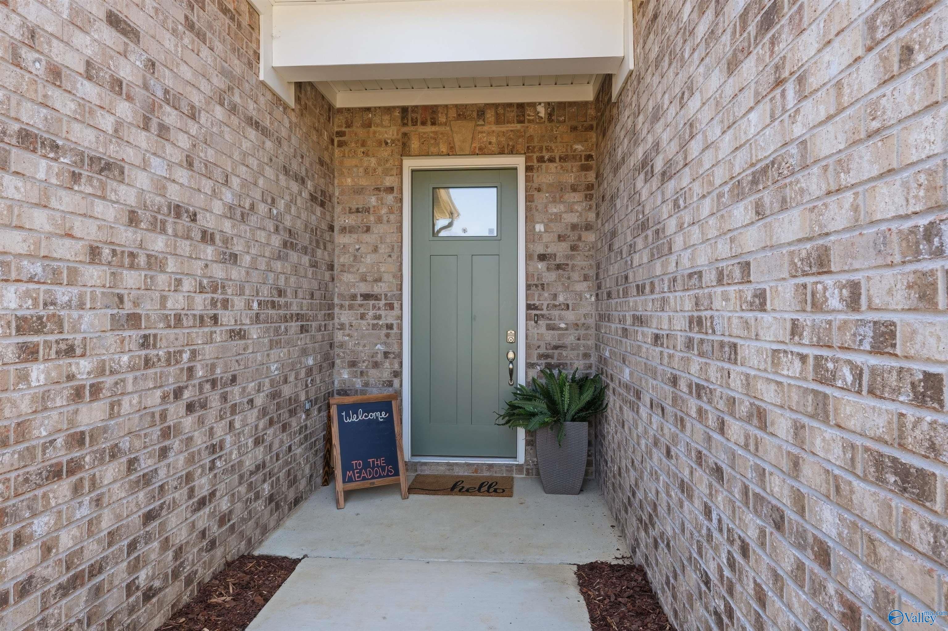 Inviting brick front entrance with sage green door, "Welcome to the Meadows" sign, hello doormat, and potted fern for Evermore Homes Oxford B in Owens Cross Roads, AL