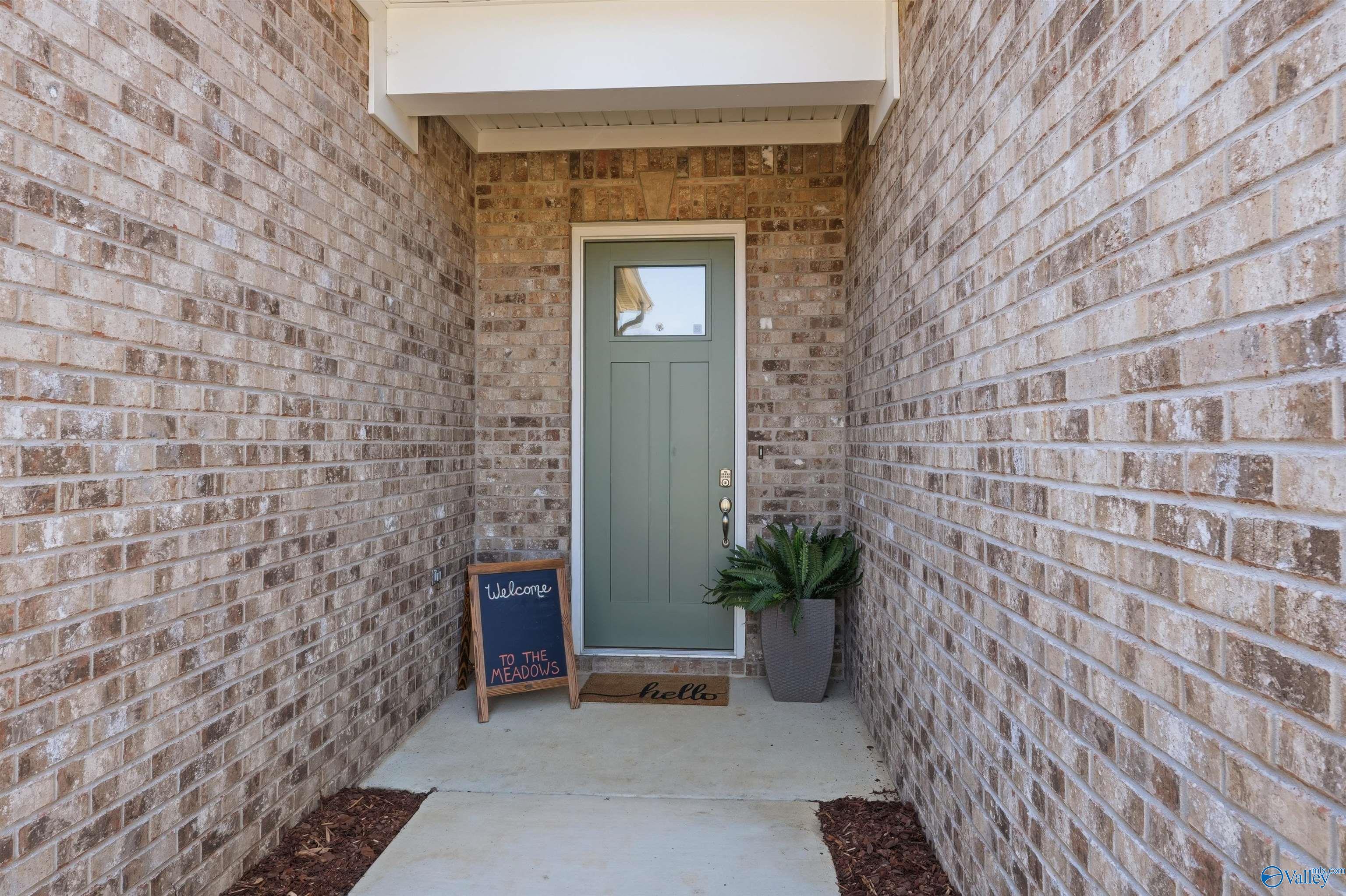 Inviting brick front entrance with sage green door, "Welcome to the Meadows" sign, hello doormat, and potted fern for Evermore Homes Oxford B in Owens Cross Roads, AL
