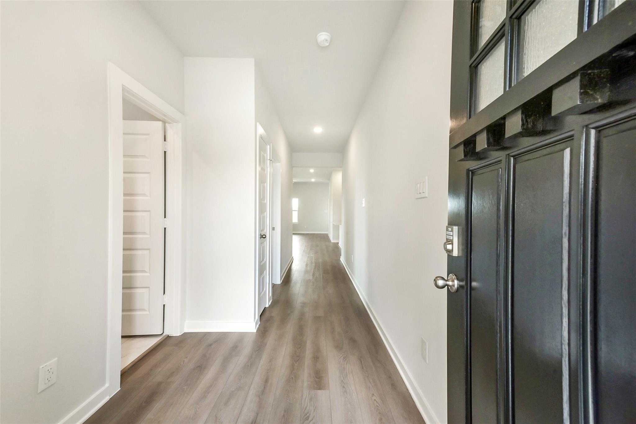 Long hallway with light wood floors, white walls, side doors, and black glass front door in Davidson Homes The Laguna C, Dayton, Texas