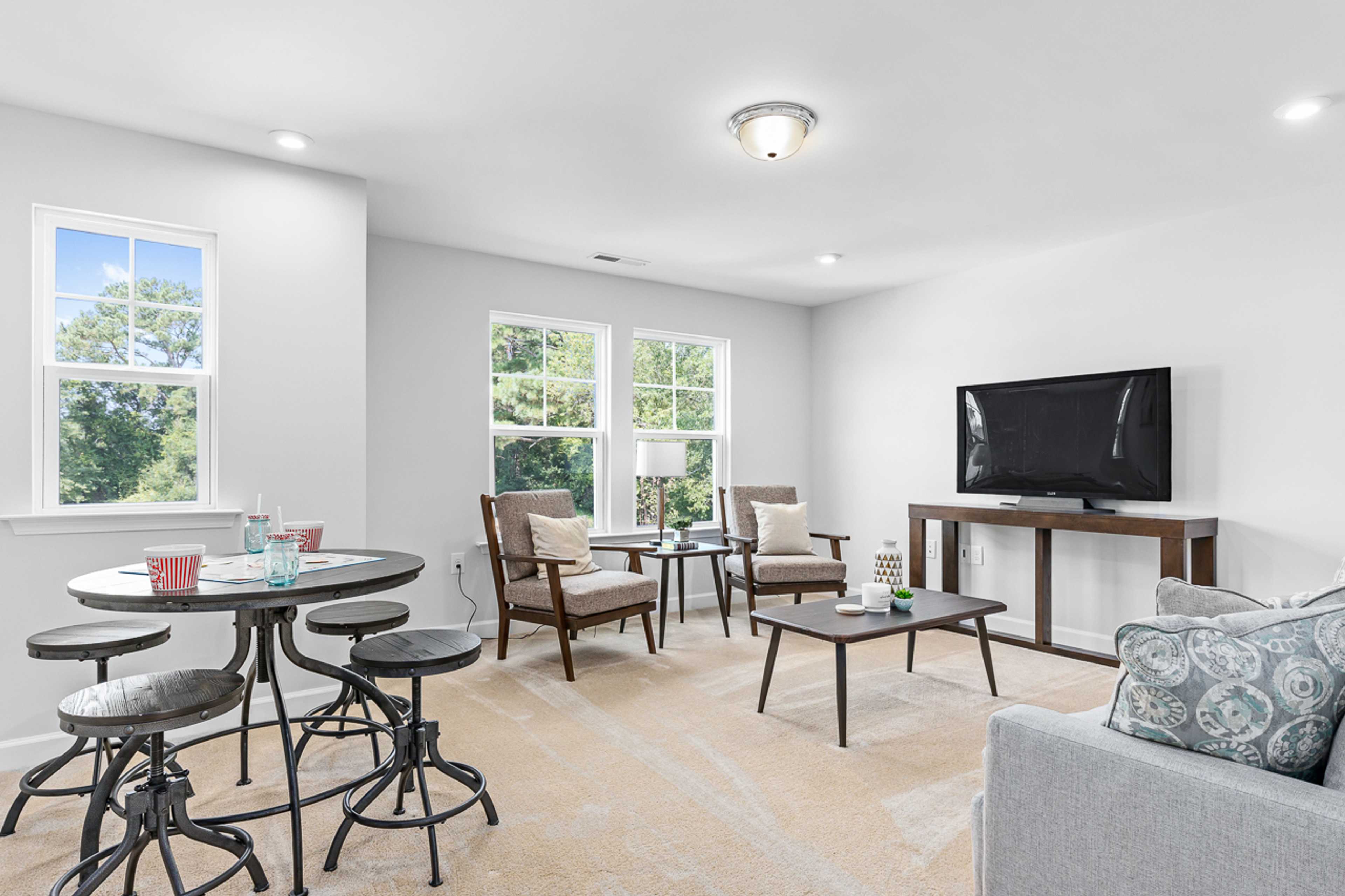 Spacious living room at Stagecoach Corner in Mebane NC with round pedestal table, mounted TV, gray sofa, armchairs, and large windows