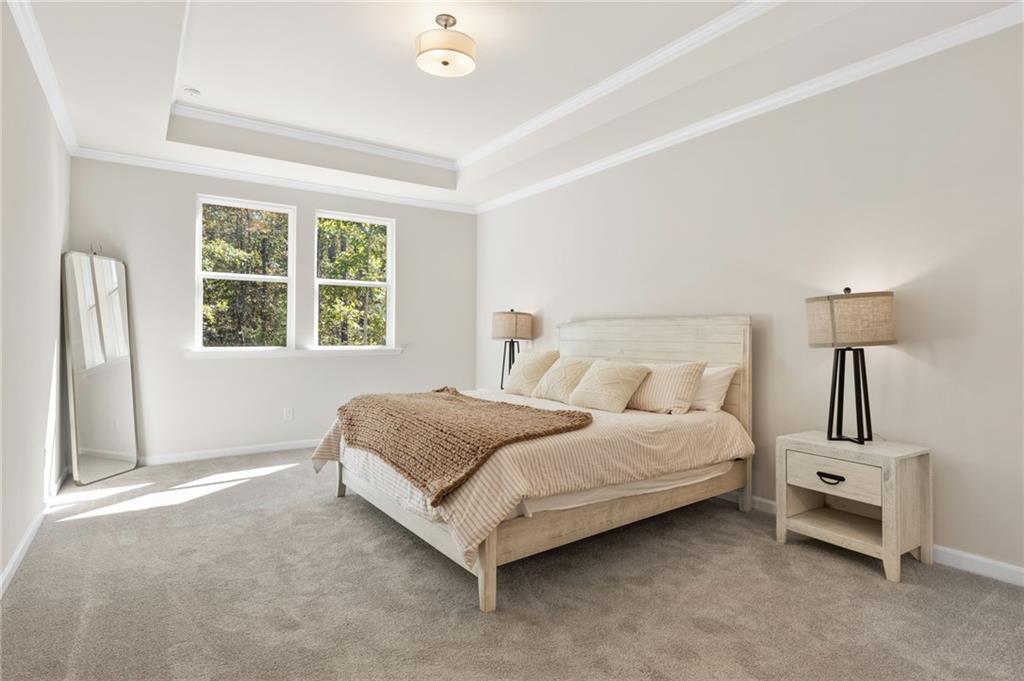 Elegant master bedroom with tray ceiling, king bed in neutral tones, and tree-lined window views in Davidson Homes The Daphne B, Loganville, GA