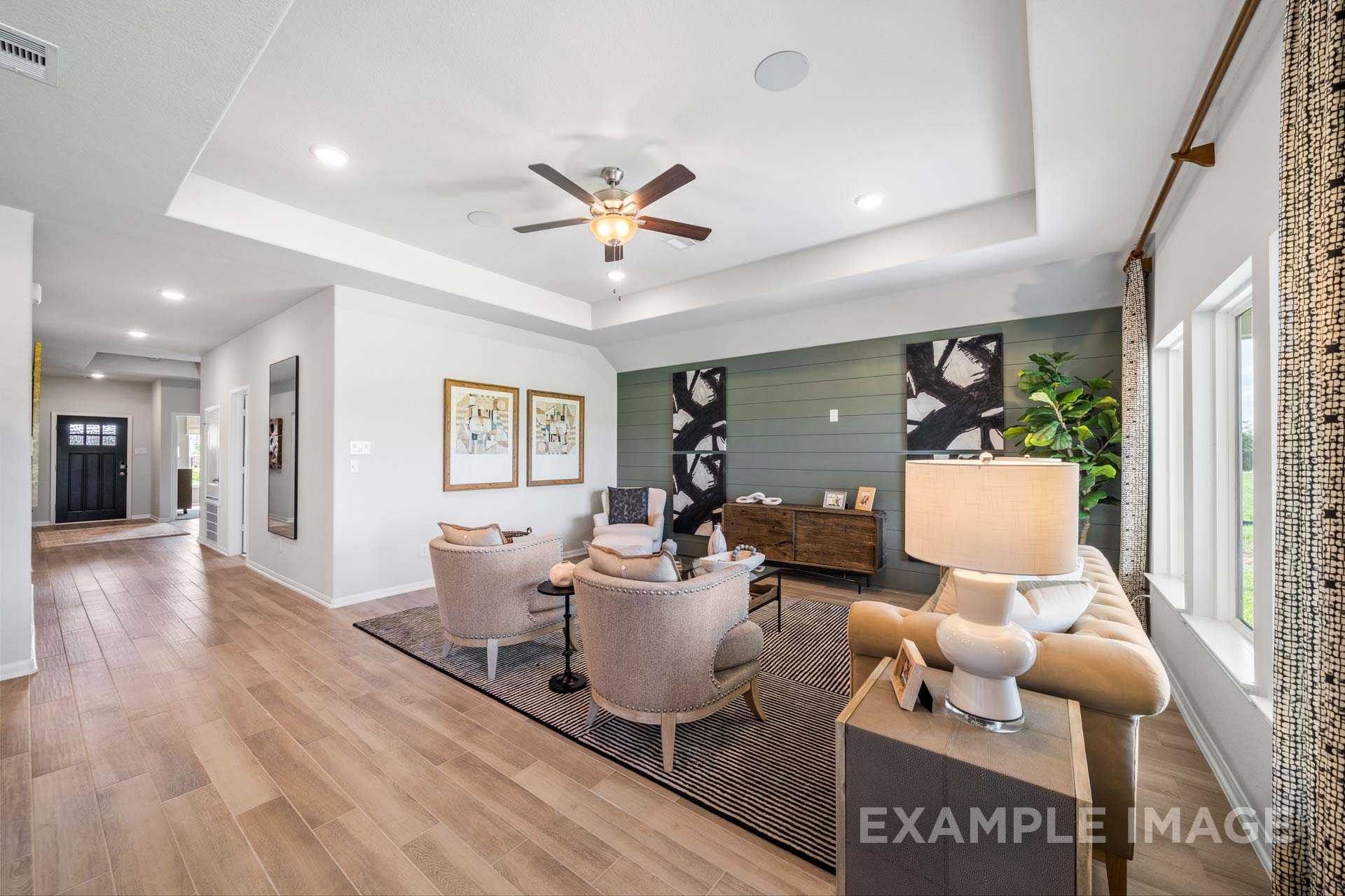 Spacious living room in The Acadia B showcasing plush armchairs, accent wall art, hardwood floors, ceiling fan, and large windows