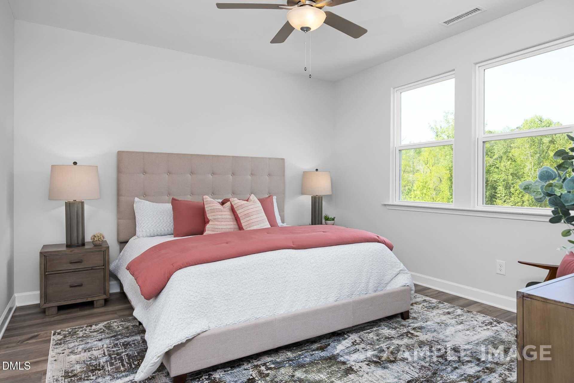 Cozy bedroom with tufted beige headboard, pink bedding, ceiling fan, and large windows overlooking trees in Davidson Homes The Carter C, Lillington, NC