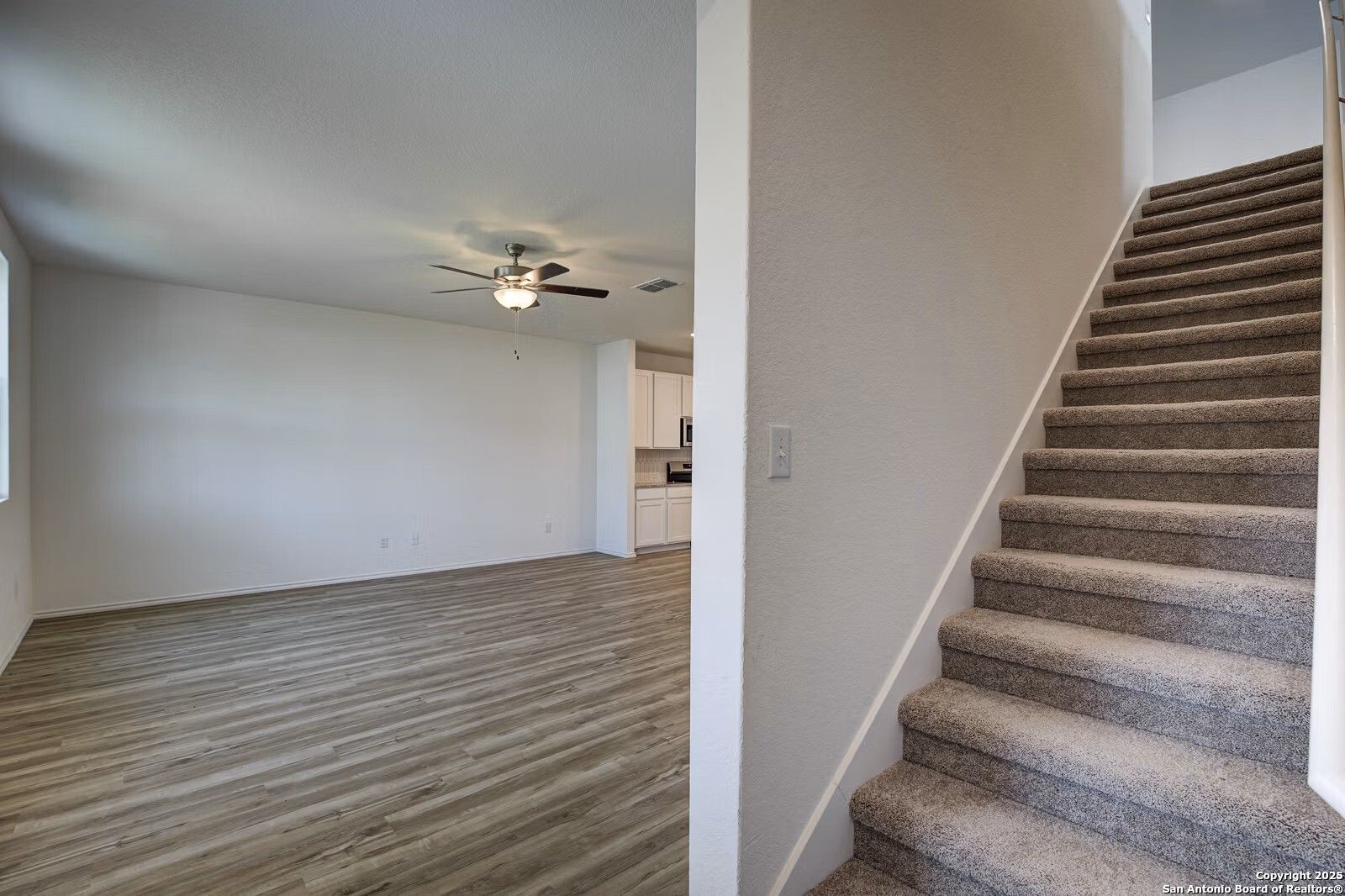 Spacious empty living room with luxury vinyl plank floors, ceiling fan, and open kitchen view beside carpeted staircase in Davidson Homes The Douglas B, Seguin, Texas