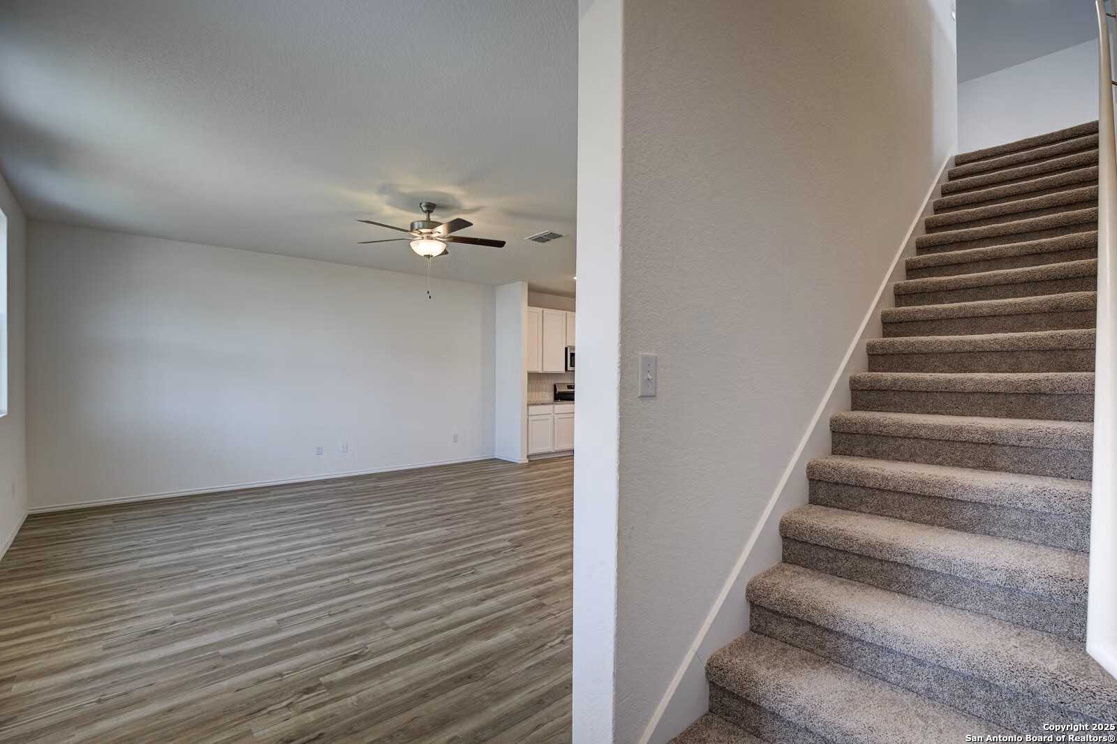 Spacious empty living room with luxury vinyl plank floors, ceiling fan, and open kitchen view beside carpeted staircase in Davidson Homes The Douglas B, Seguin, Texas