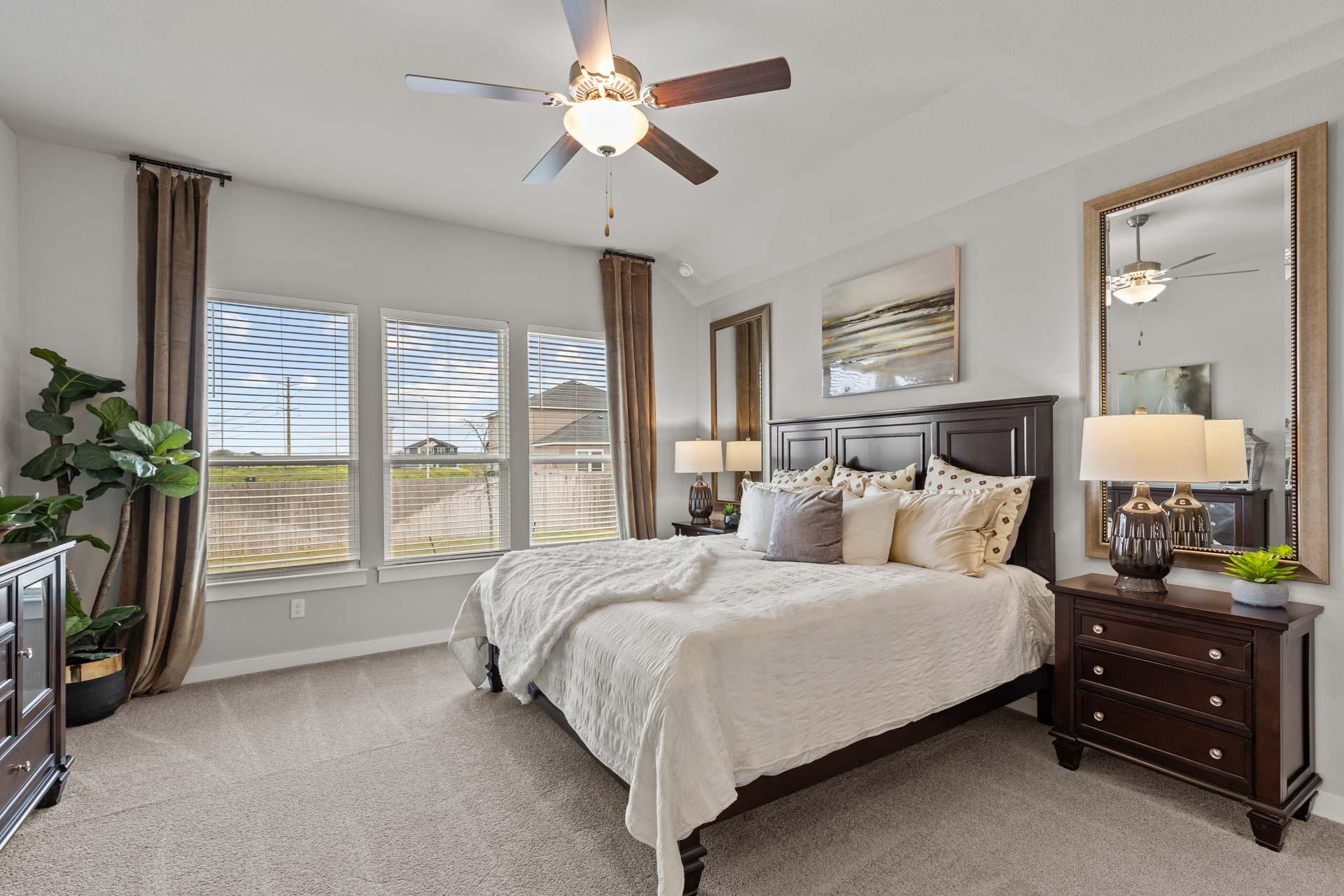 Spacious master bedroom in The Collin A featuring king bed with neutral bedding, large windows, ceiling fan, and potted plants