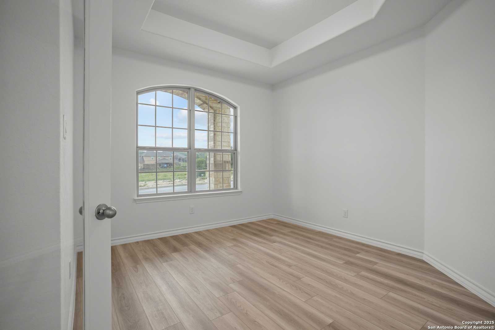 Bright bedroom with arched window, tray ceiling, and hardwood floors in Davidson Homes The Summerlin B, Castroville, Texas