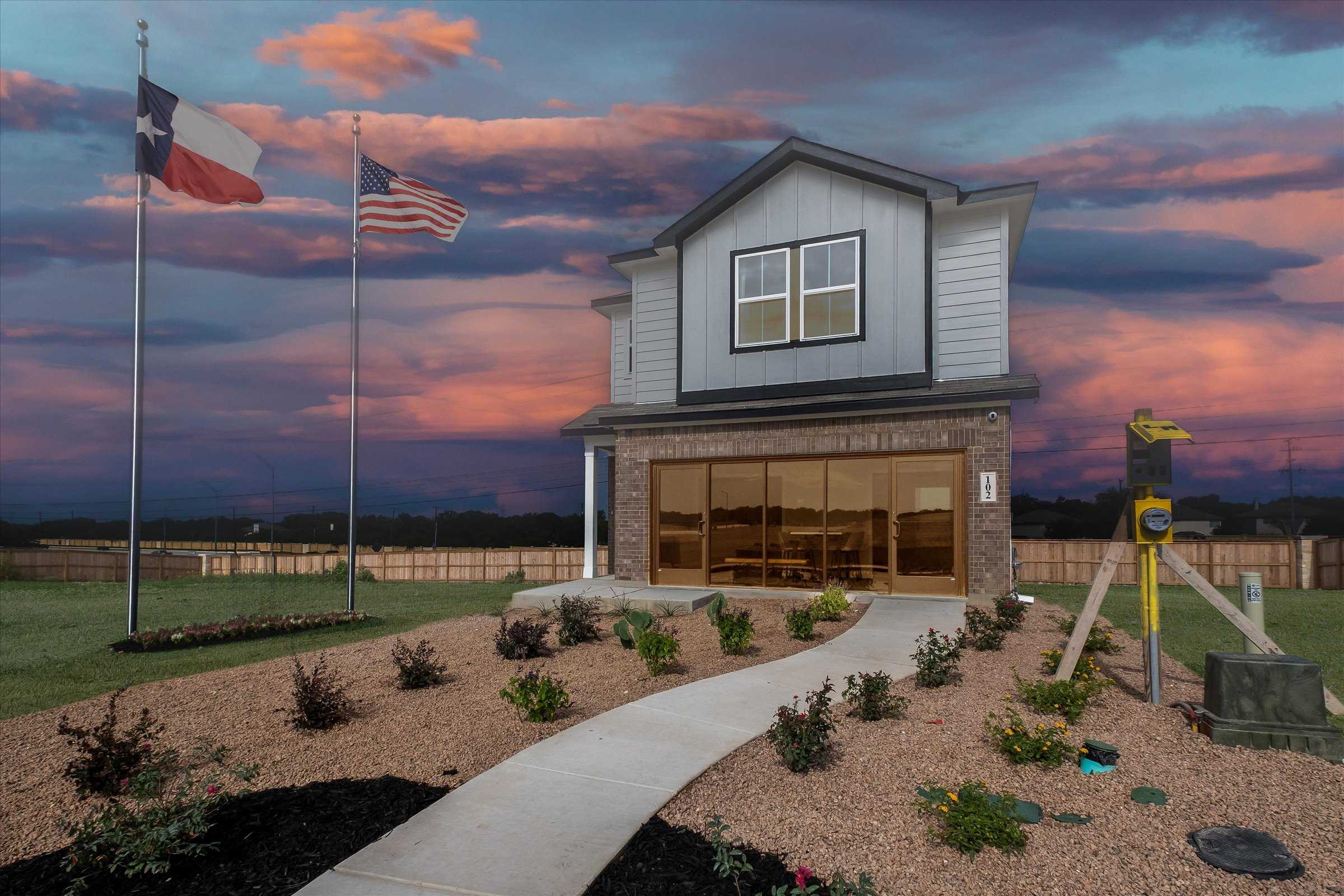 Two-story Florence C home by Davidson Homes in Meadows at Oak Creek, San Antonio, with gray siding, 2-car garage, flags, and sunset landscaping
