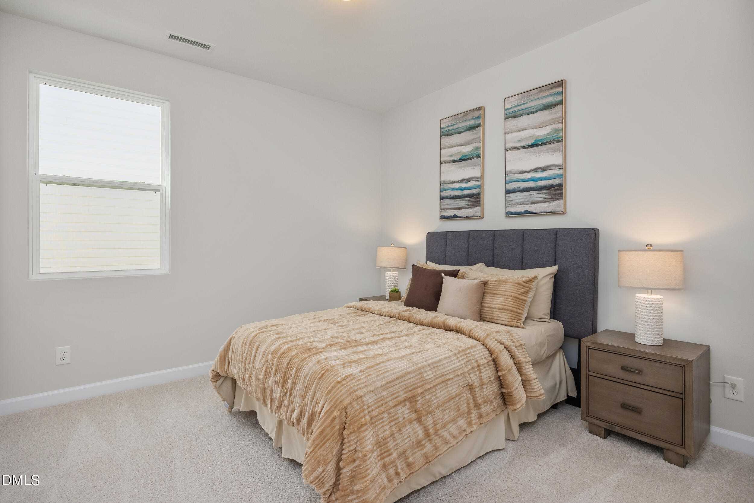 Serene secondary bedroom with beige bedding, tufted headboard, lamps, abstract art, and large window in Davidson Homes The Preston C, Lillington, NC