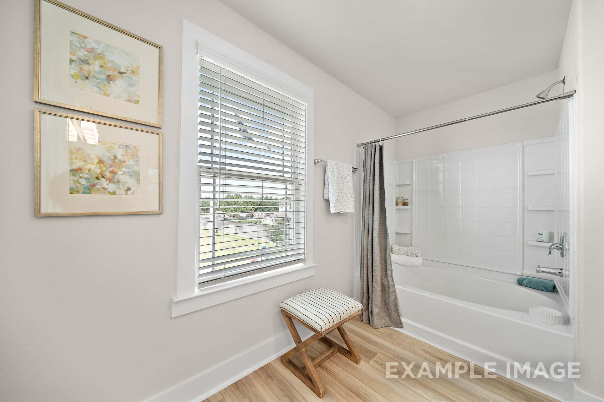 Spacious master bathroom in The Lily A with white tiled shower, soaking tub, window blinds, and artwork accents