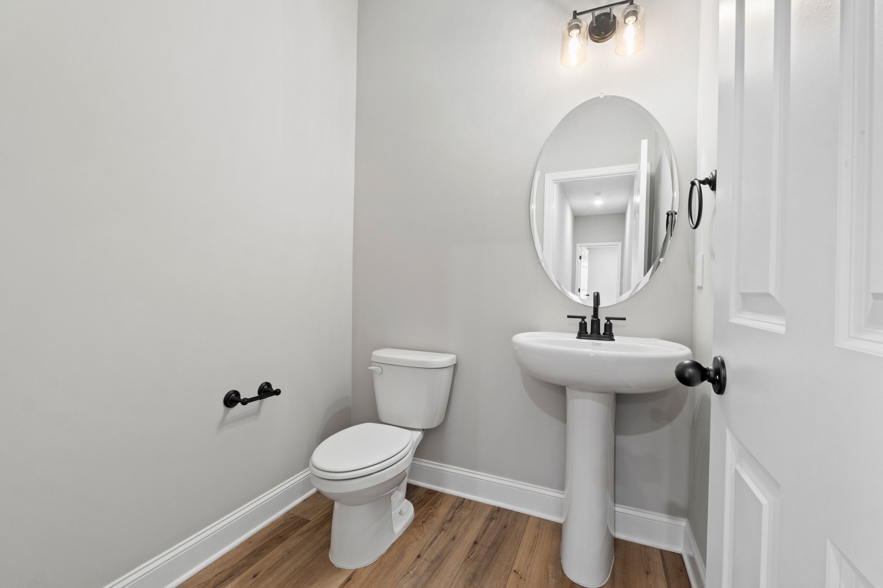 Modern powder room in The Valencia: pedestal sink, white toilet, round mirror, gray walls, hardwood floors