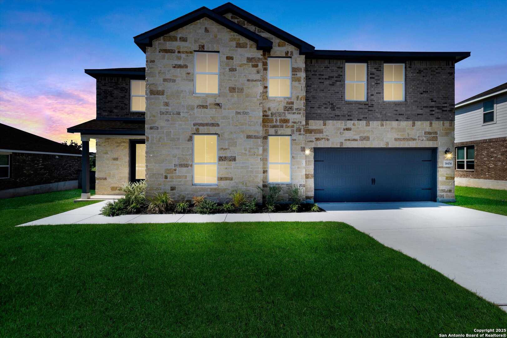 Modern two-story stone-brick home with lit windows, two-car garage, and landscaped yard at dusk in Ladera, San Antonio, Texas