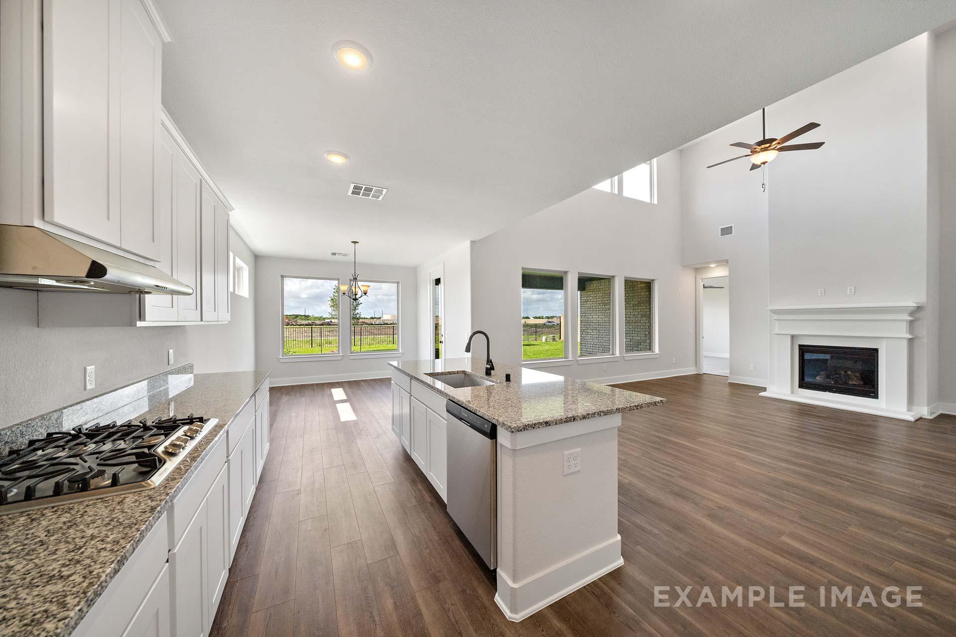 Spacious open-concept kitchen in The Victoria A with white shaker cabinets, granite island, gas range, and adjacent living area featuring fireplace