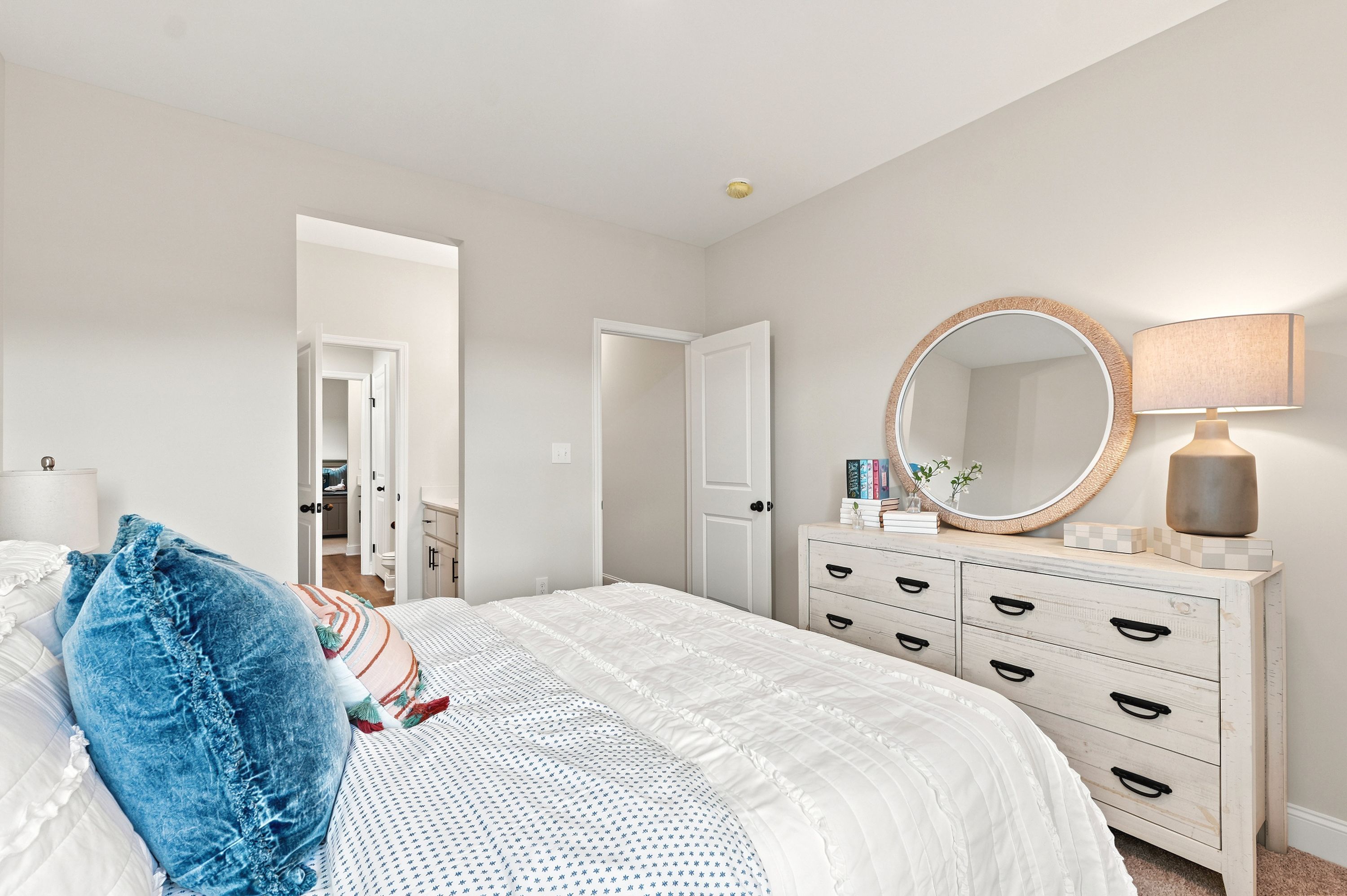Cozy bedroom at Riverton Preserve in Huntsville, AL by Davidson Homes featuring a white dresser, round mirror, and bed.