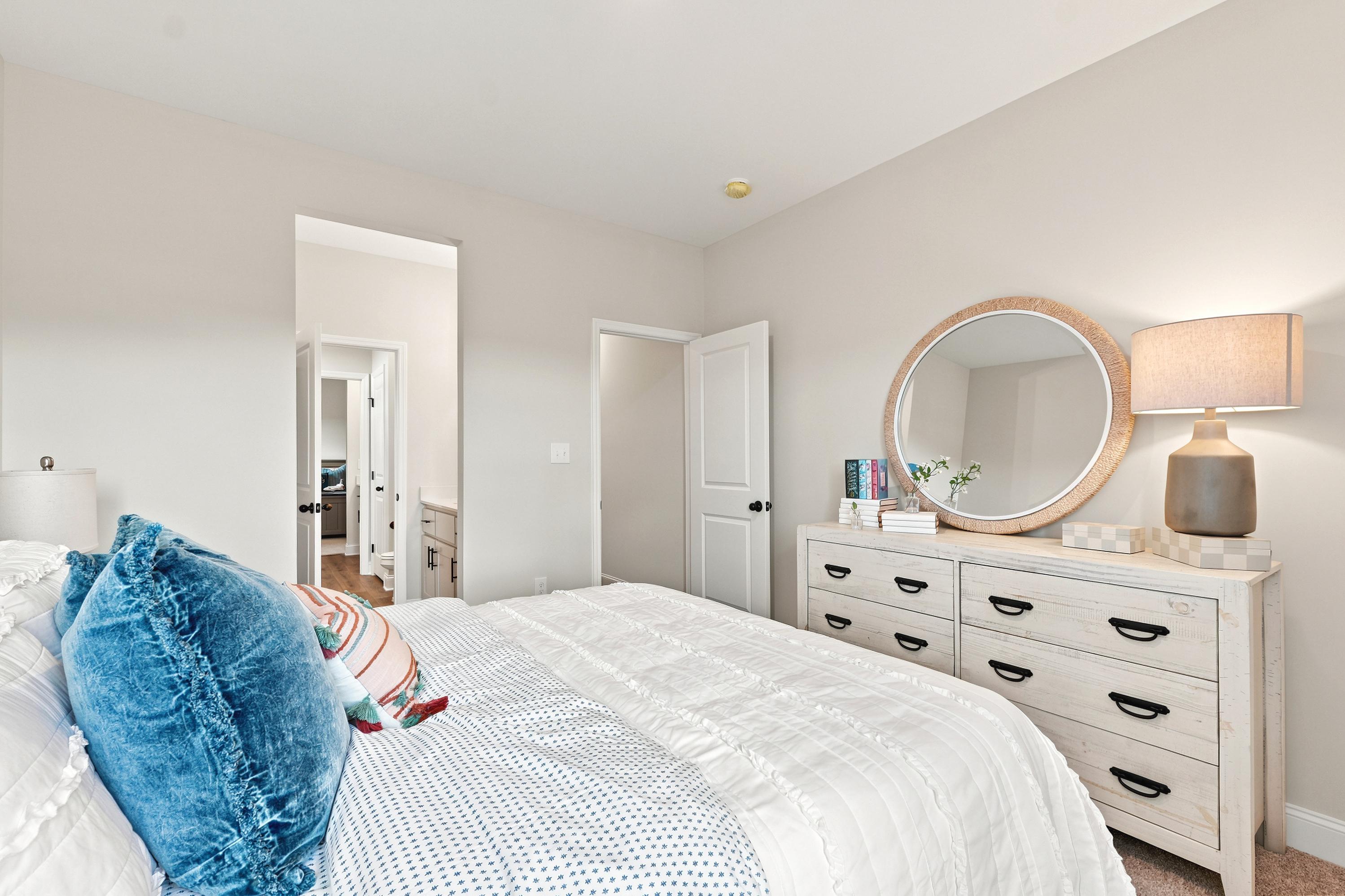 Cozy bedroom at Riverton Preserve in Huntsville, AL by Davidson Homes featuring a white dresser, round mirror, and bed.