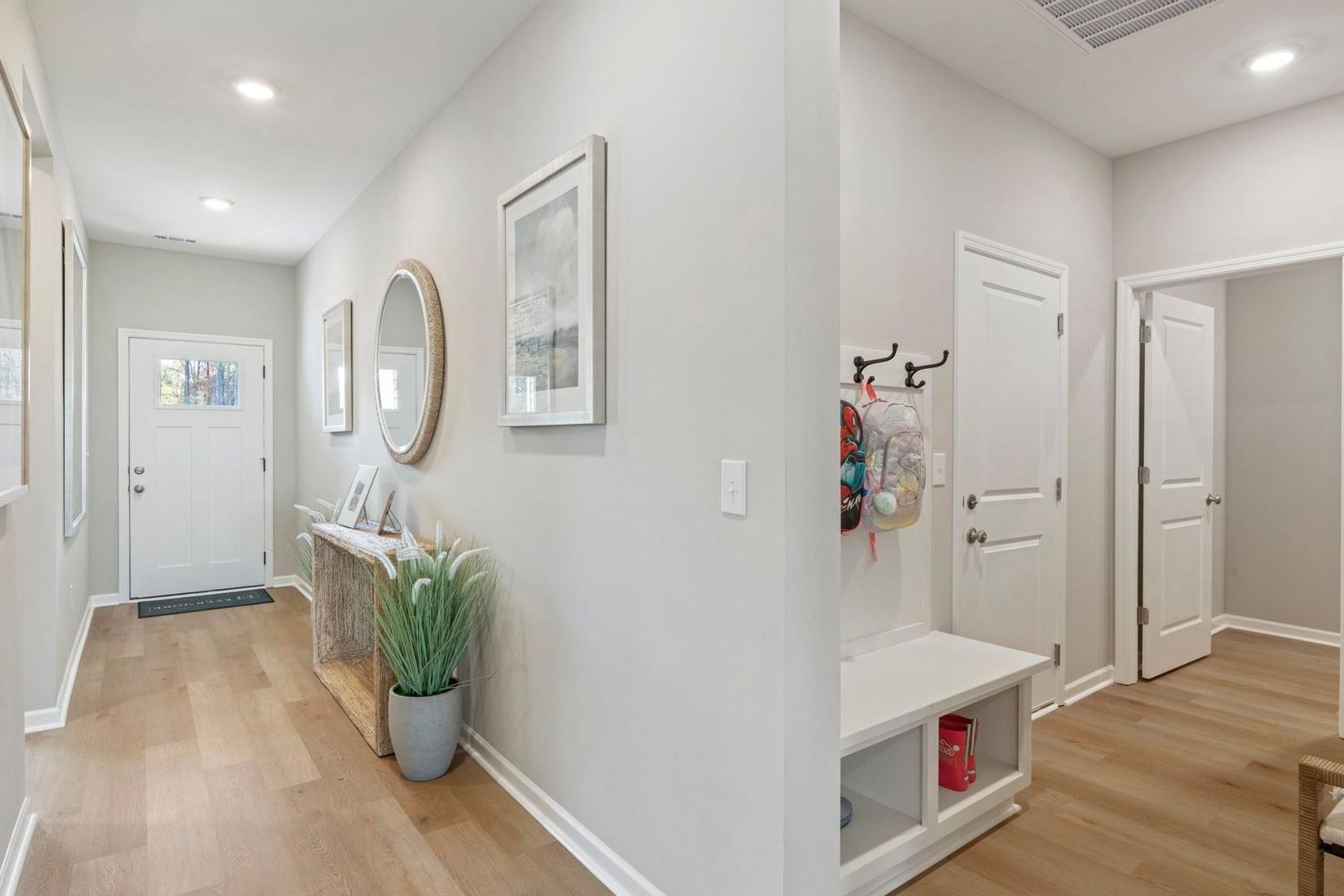 Welcoming entryway in The Phoenix A by Davidson Homes featuring white walls, wood floors, console table with plants, round mirror, and built-in bench with hooks