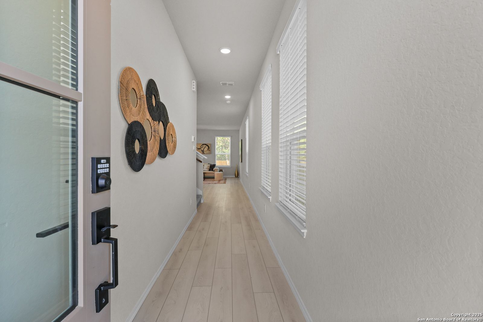 Elegant light gray hallway with wooden circular wall art, blinds-covered windows, and beige plank flooring in Davidson Homes The Charlotte B, San Antonio