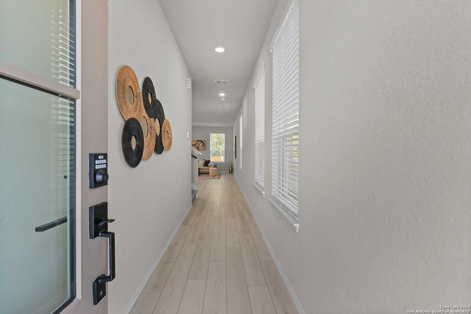 Elegant light gray hallway with wooden circular wall art, blinds-covered windows, and beige plank flooring in Davidson Homes The Charlotte B, San Antonio