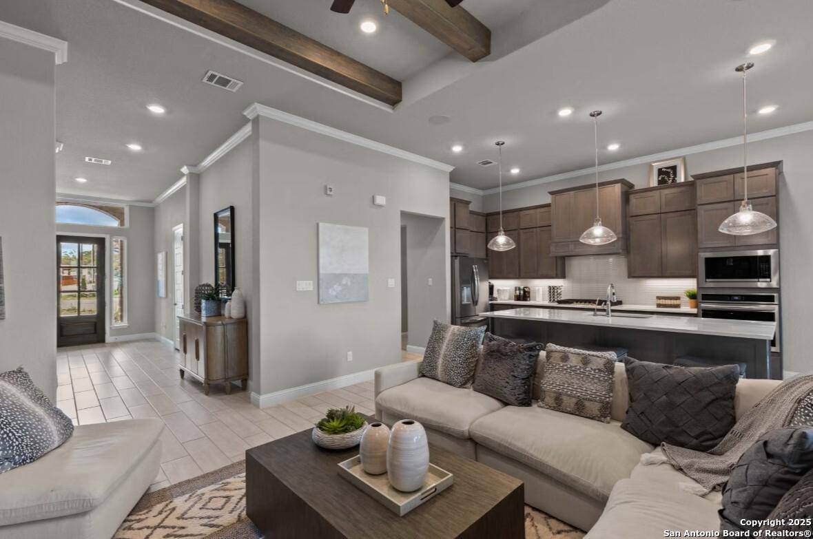 Open-concept living room with gray sectional sofa, wooden coffee table, and adjacent kitchen featuring brown cabinets, island, pendant lights in The Garner B, Ladera, San Antonio