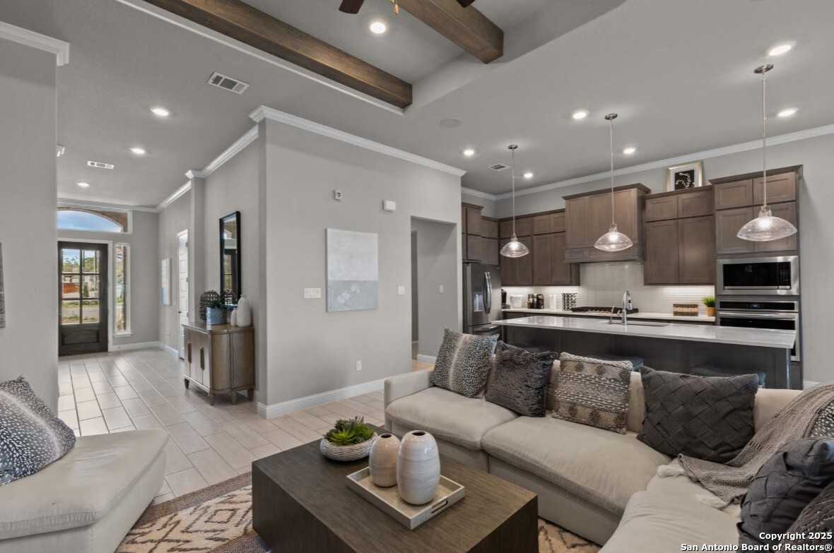 Open-concept living room with gray sectional sofa, wooden beams, and adjacent kitchen featuring brown cabinets and island in Davidson Homes The Garner B, Ladera, San Antonio