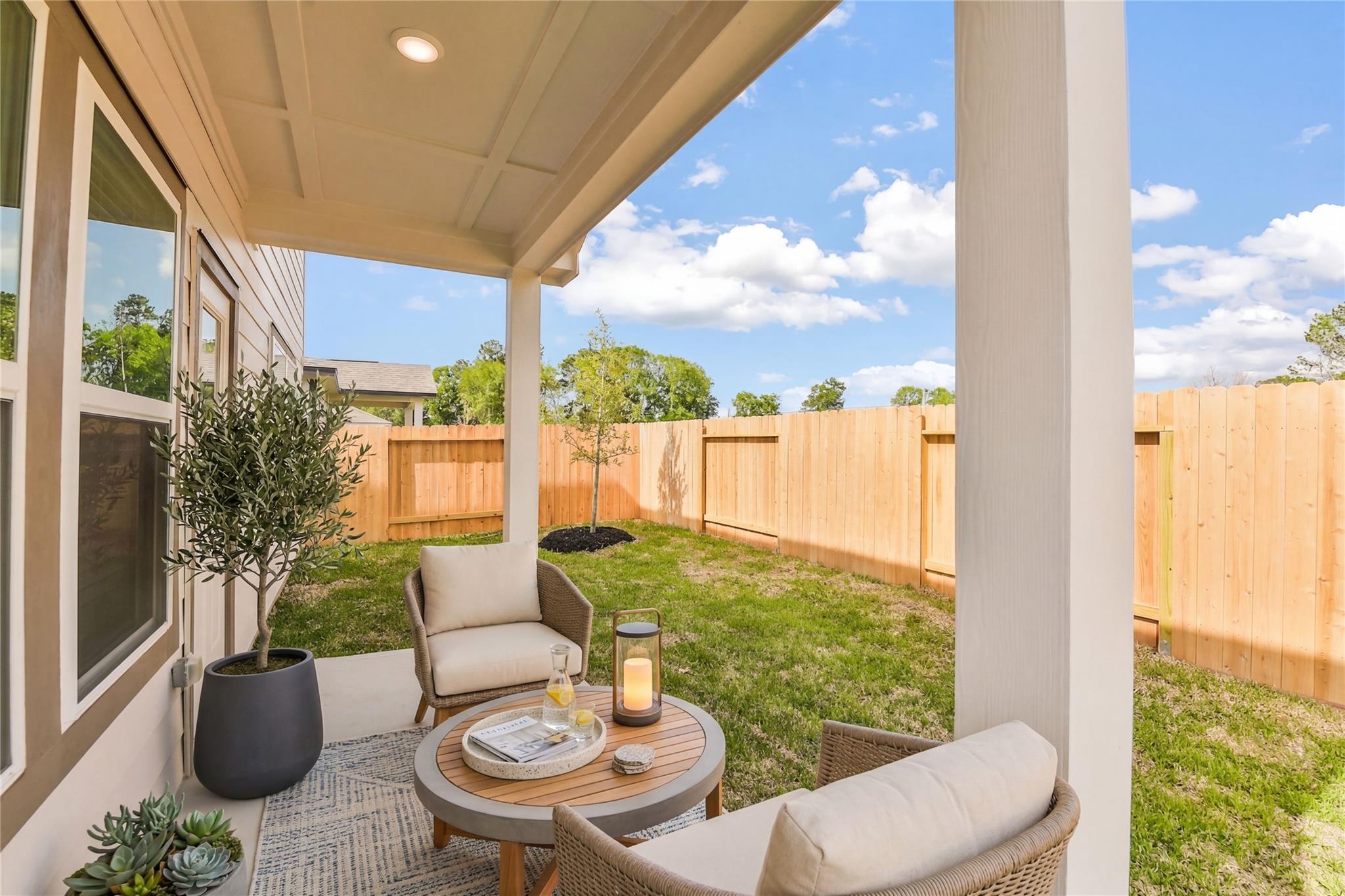 Cozy covered patio with wicker chairs, round table, potted olive tree, and lush green lawn in fenced backyard of Davidson Homes San Marcos E, Conroe Texas
