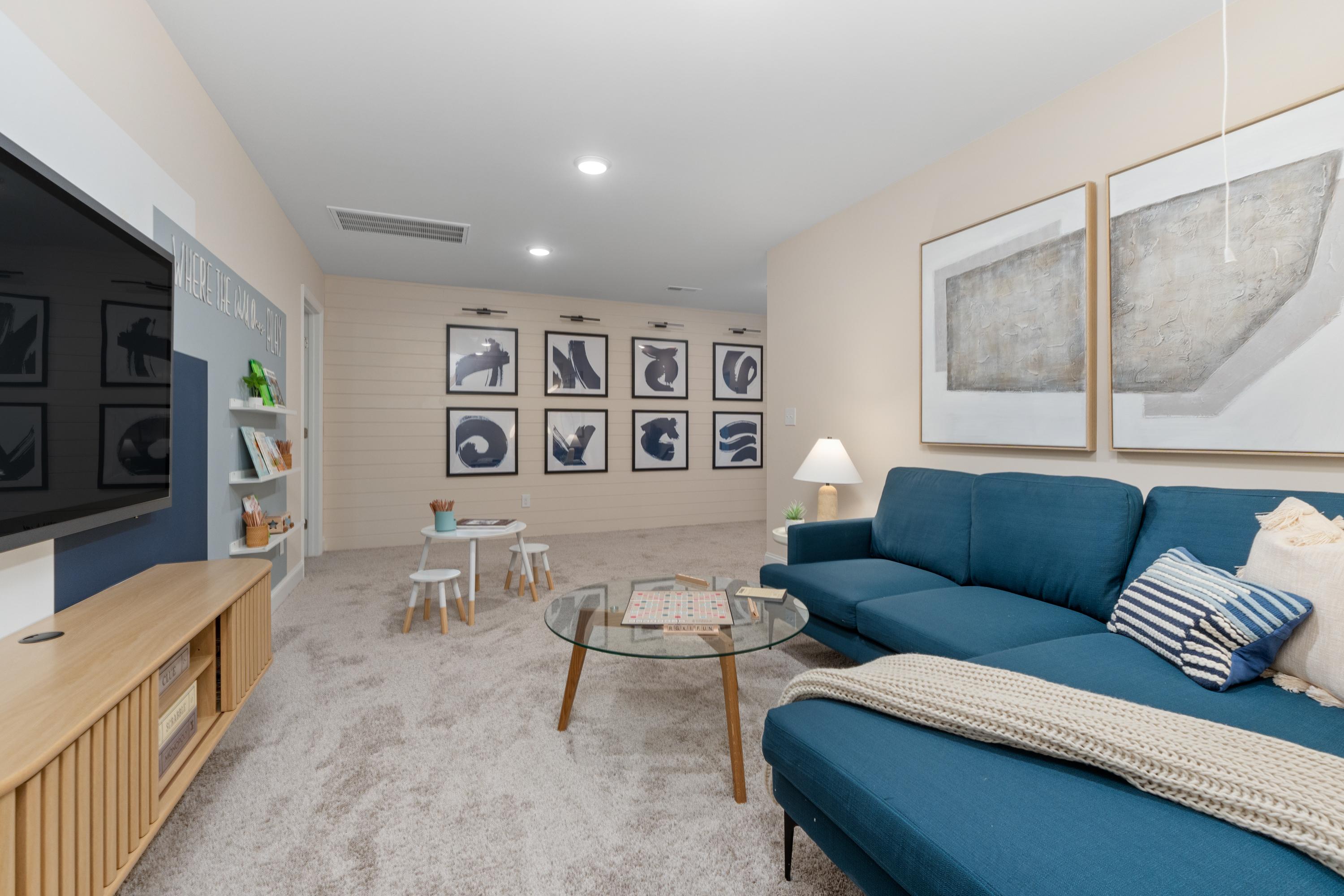 Spacious bonus room in Pavilion Huntsville AL with blue sectional sofa, TV on media wall, kids' play table, abstract art, and hardwood accents